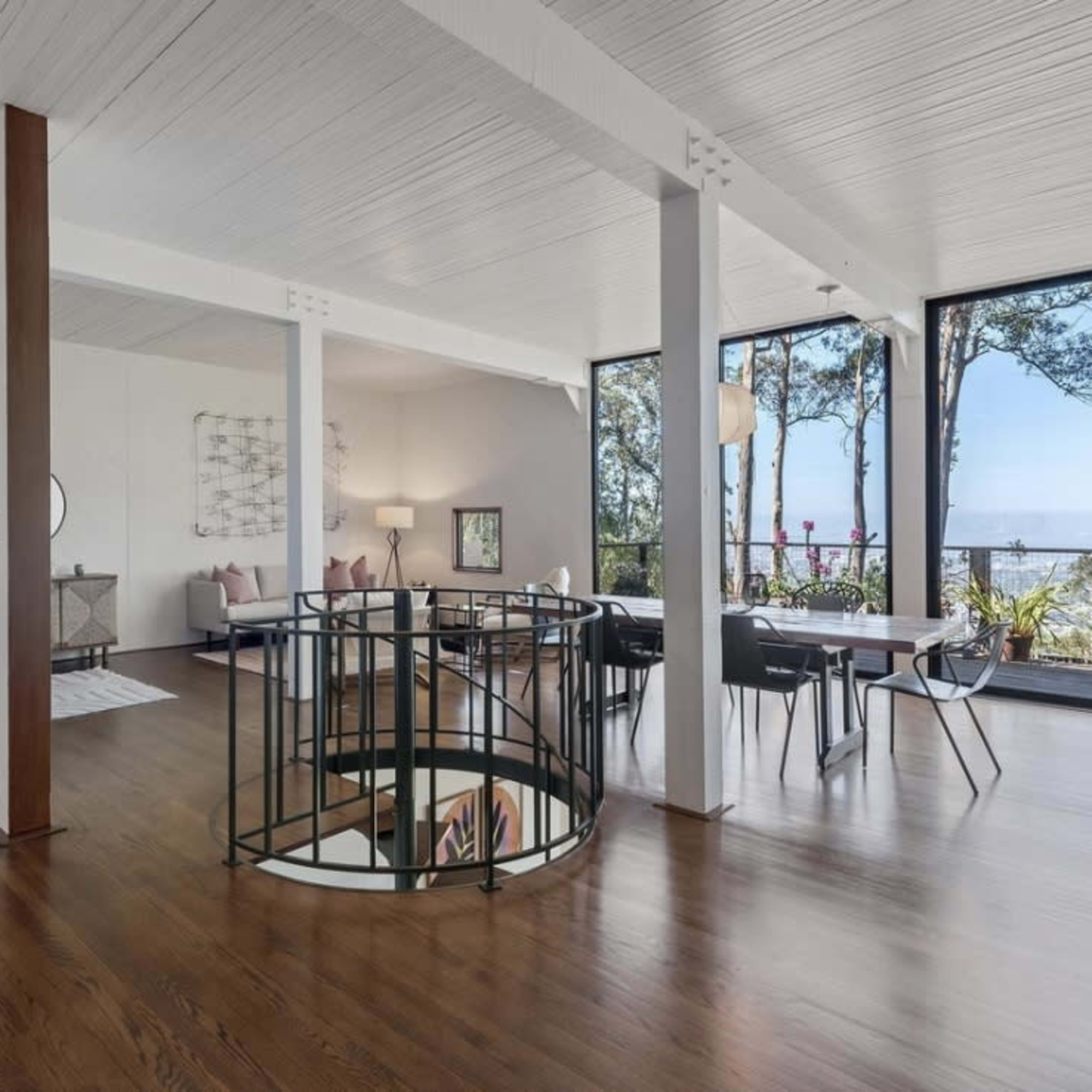 A modern, open-concept living area with large windows providing views of trees and a body of water, featuring wooden flooring and a curved staircase.
