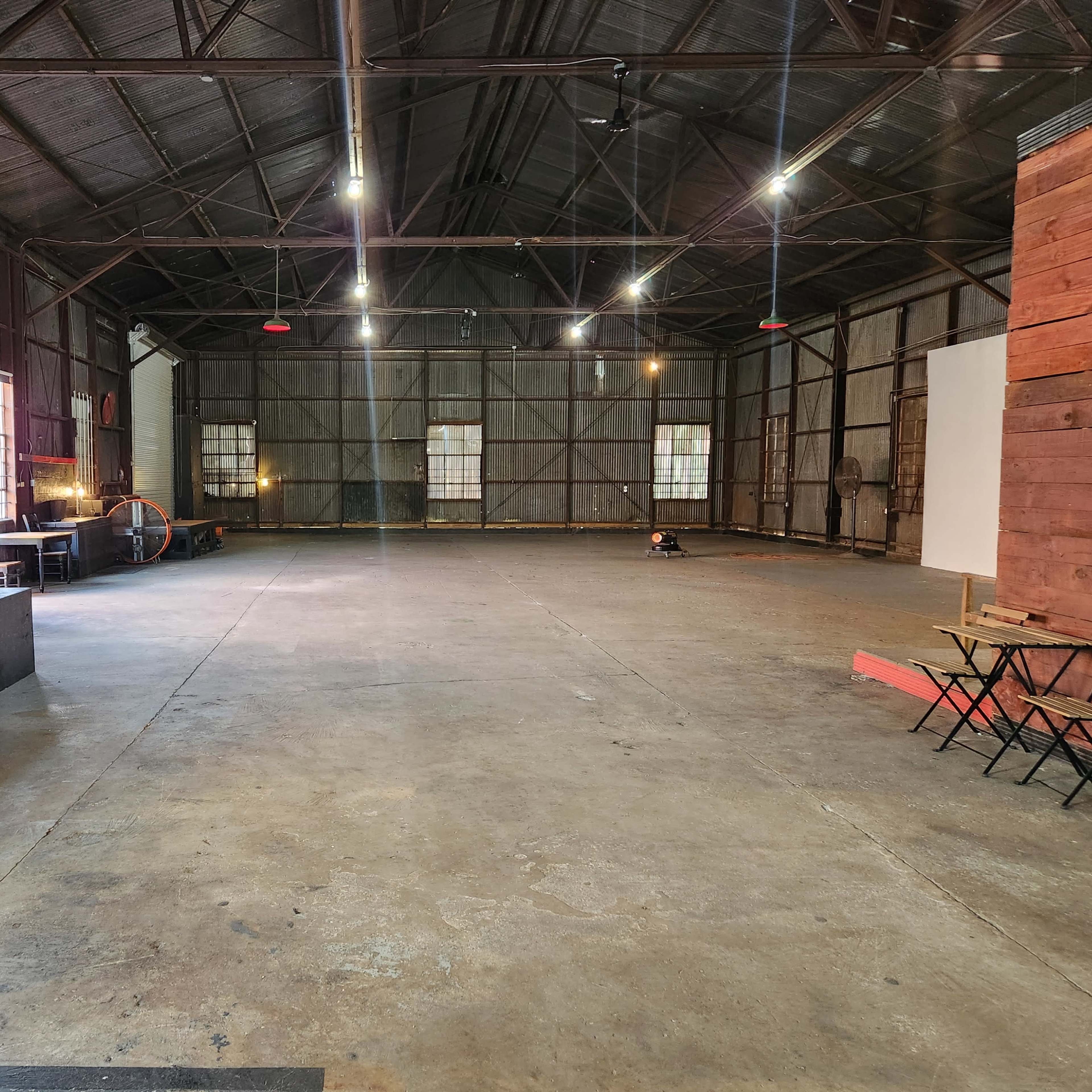 A large, empty industrial space with high ceilings, exposed metal beams, and concrete flooring, featuring wooden structures and furniture on one side.