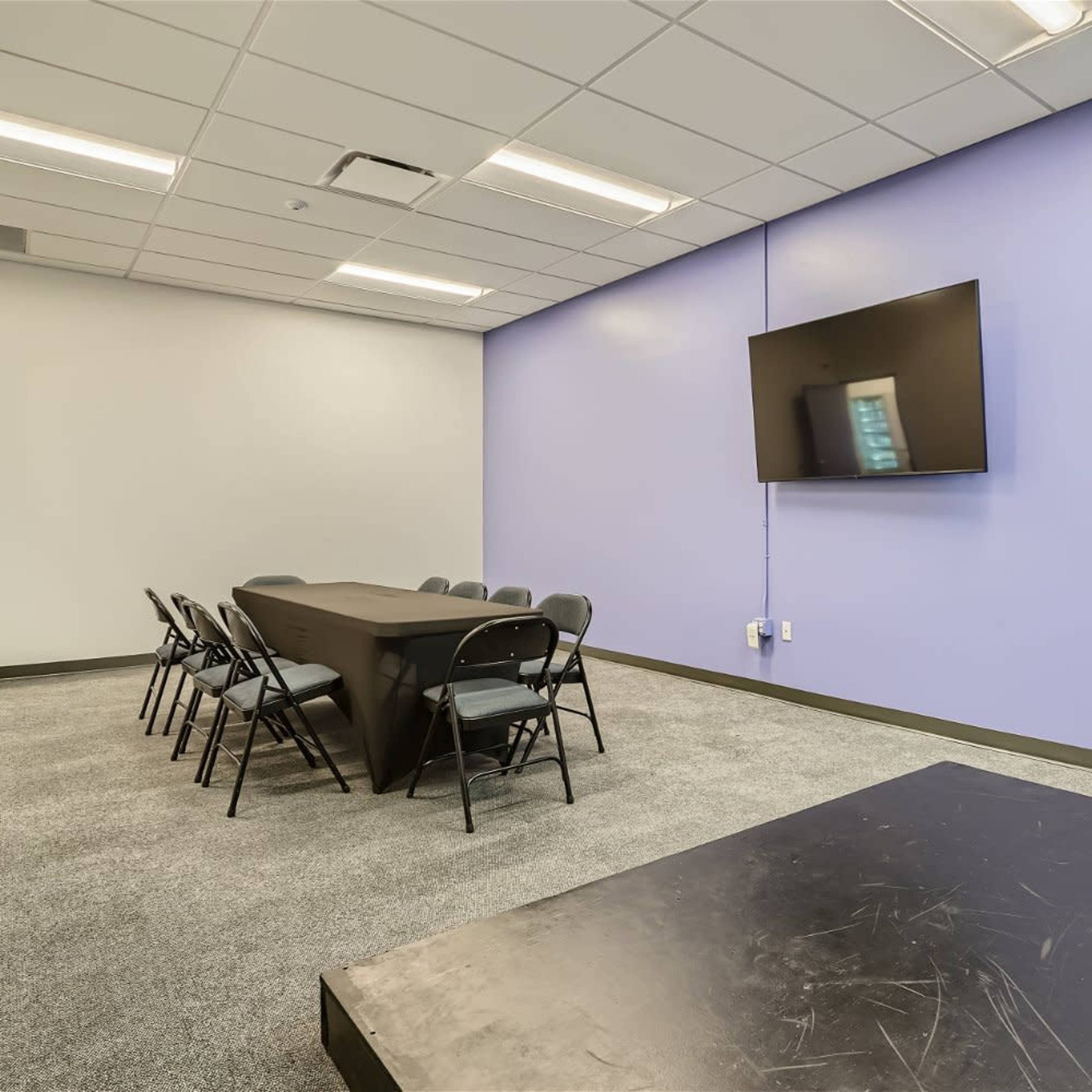 A meeting room features a black table surrounded by seven folding chairs, a large flat-screen television mounted on a purple wall, and gray carpeting.