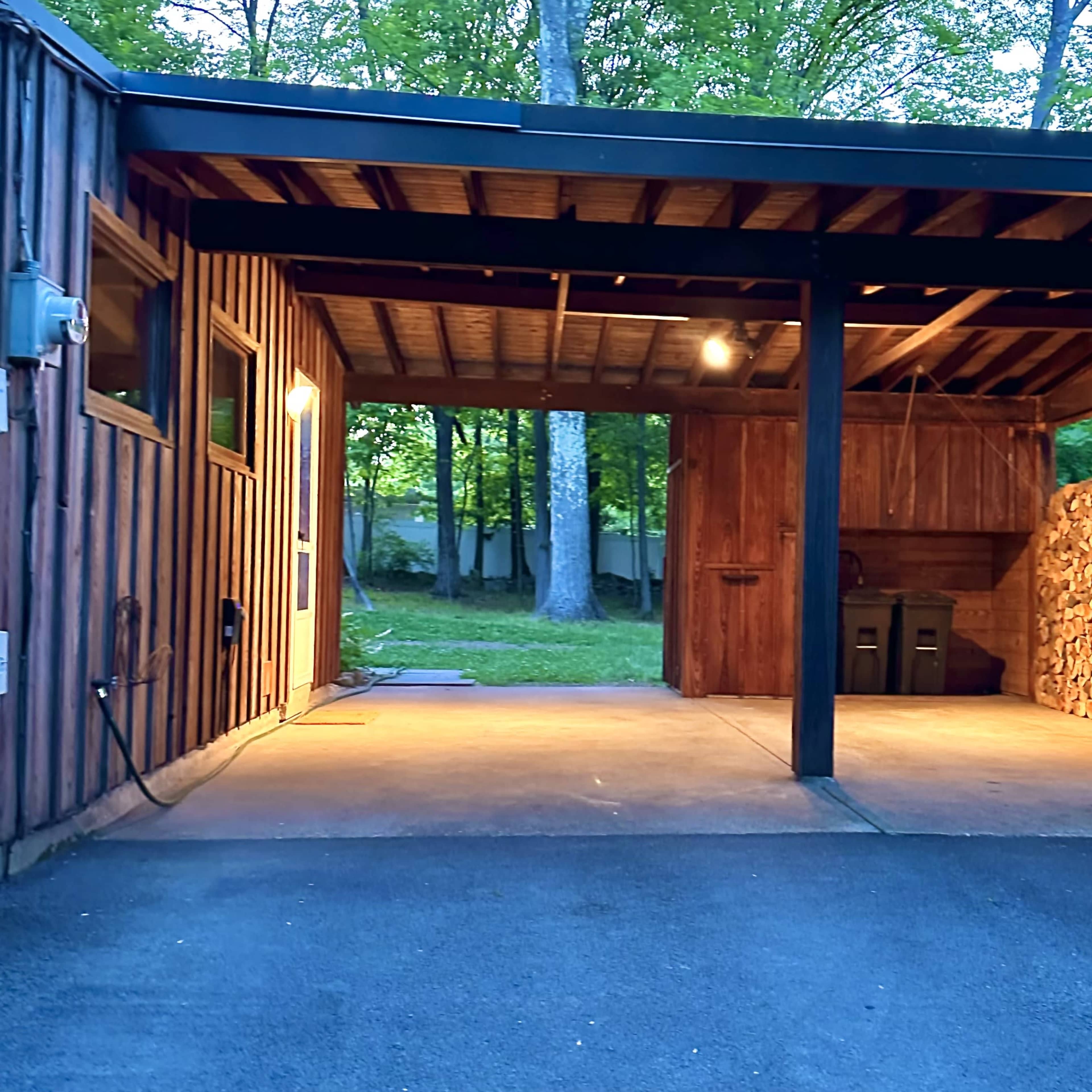 A carport with wooden walls and an outdoor storage area stacked with firewood, illuminated by a ceiling light.