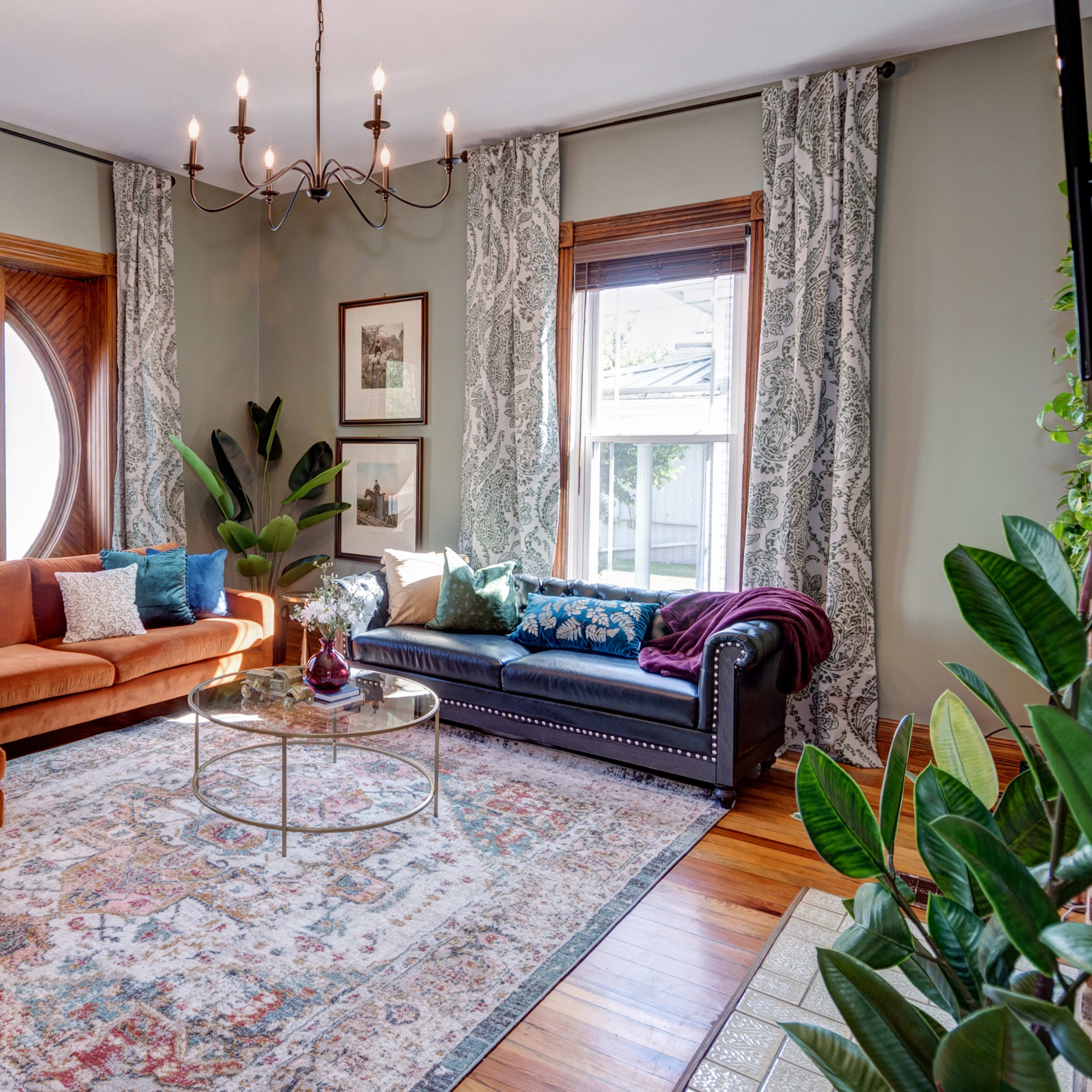 A living room with a brown sectional sofa, a black sofa, a round coffee table on a patterned rug, and large windows with decorative curtains.
