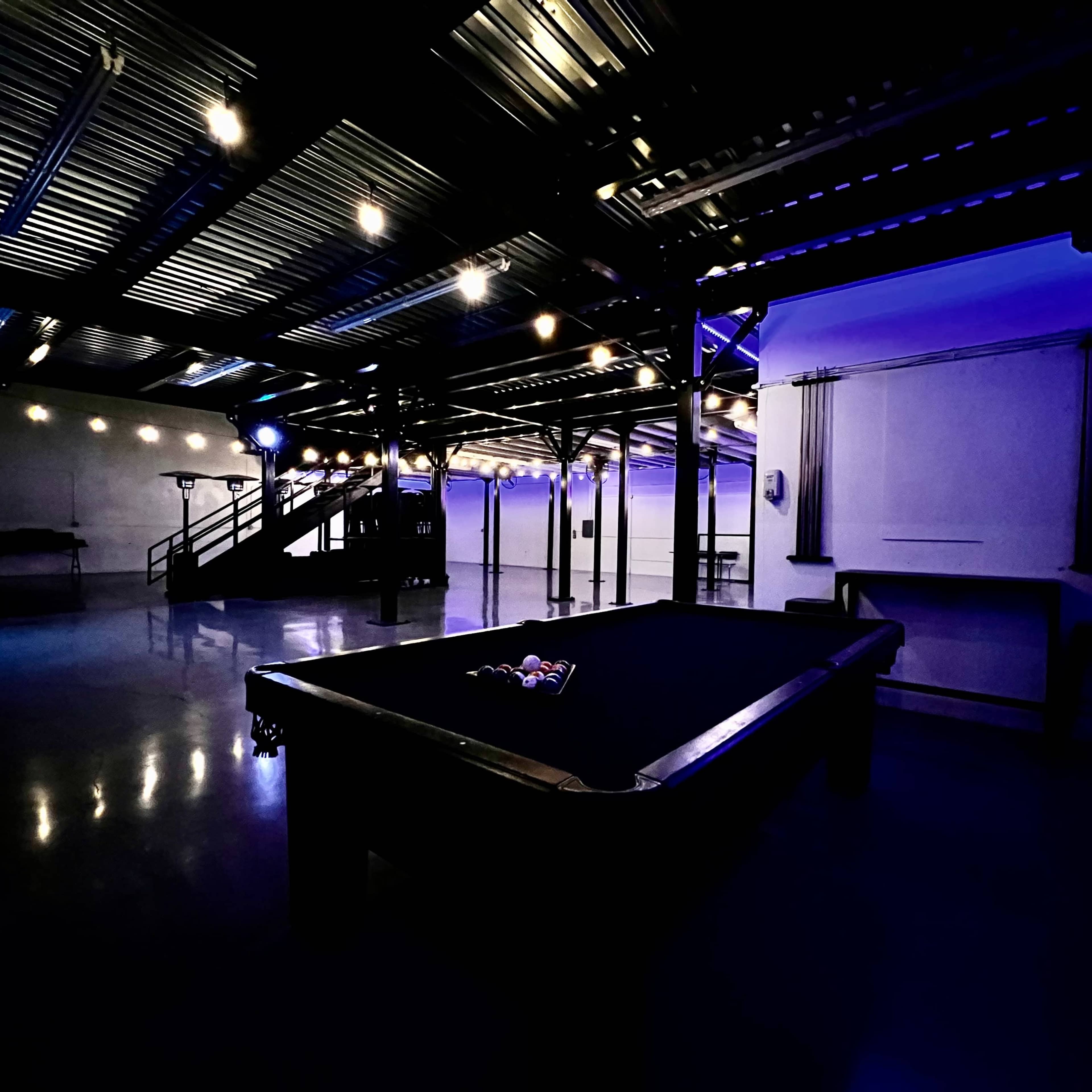 A modern event space with a wooden floor, shelving displaying bottles, and purple lighting accentuated by chandeliers.