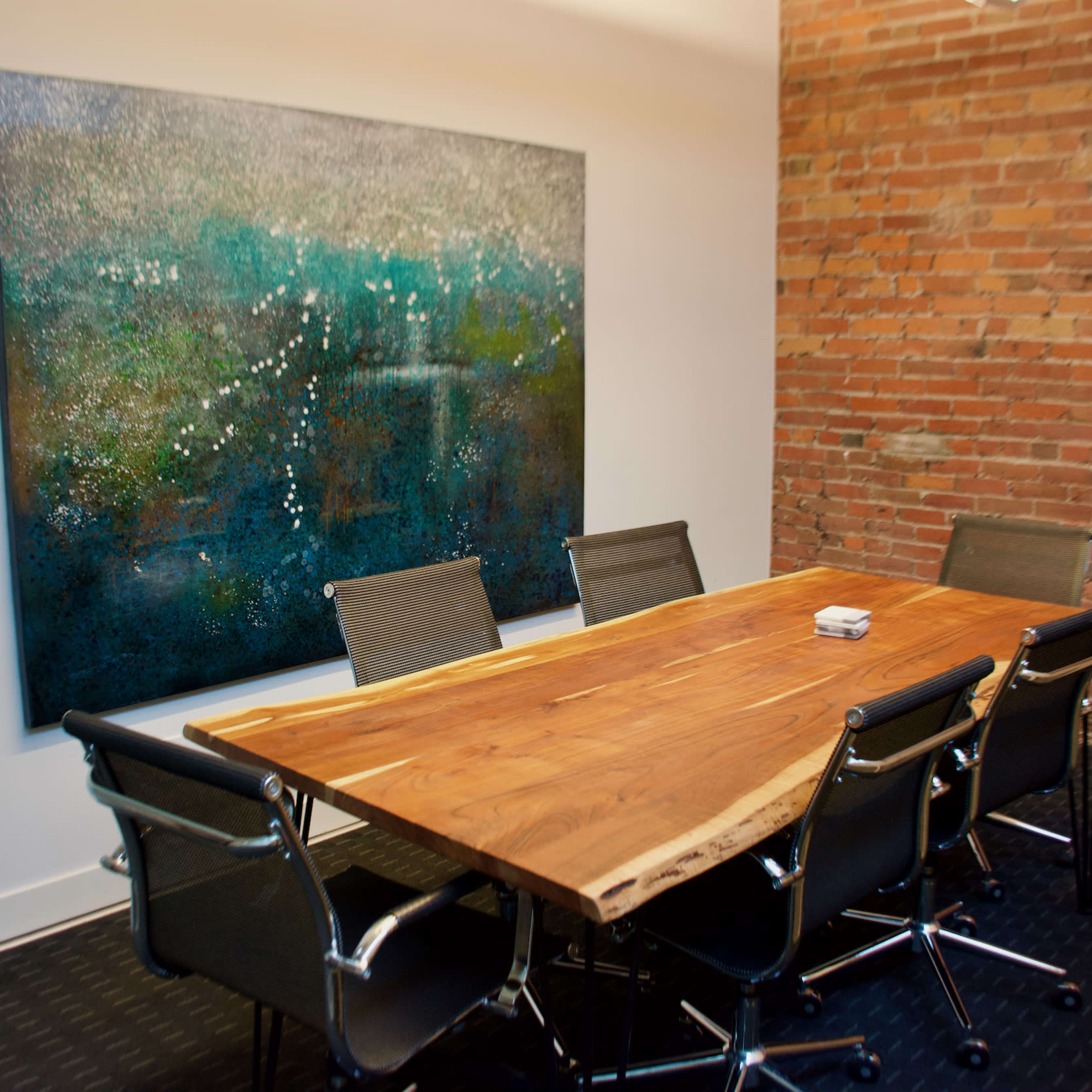 A modern conference room features a large wooden table surrounded by black mesh chairs and a colorful abstract painting on the brick wall.