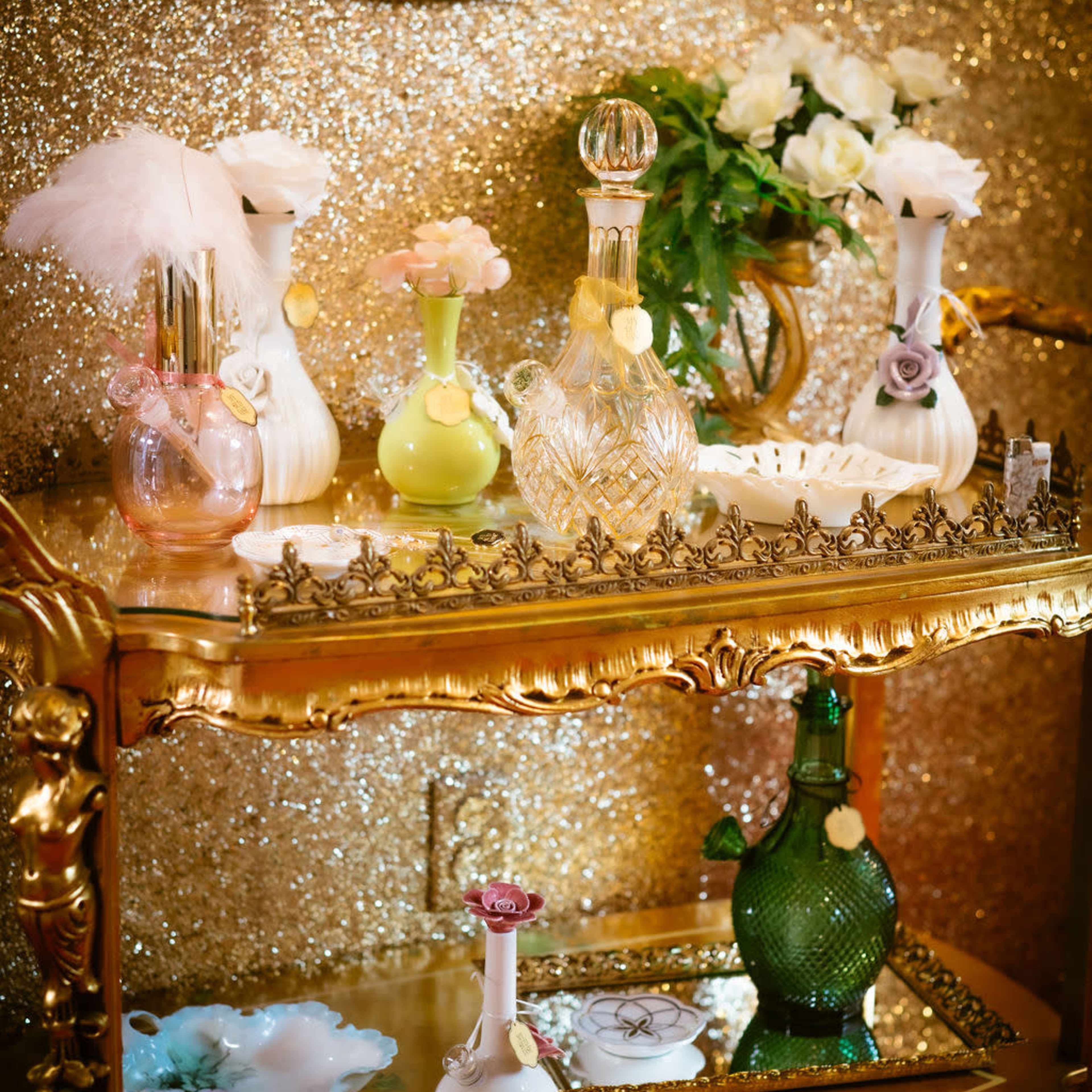 A golden serving cart displays various decorative perfume bottles and a small floral arrangement against a glittering background.