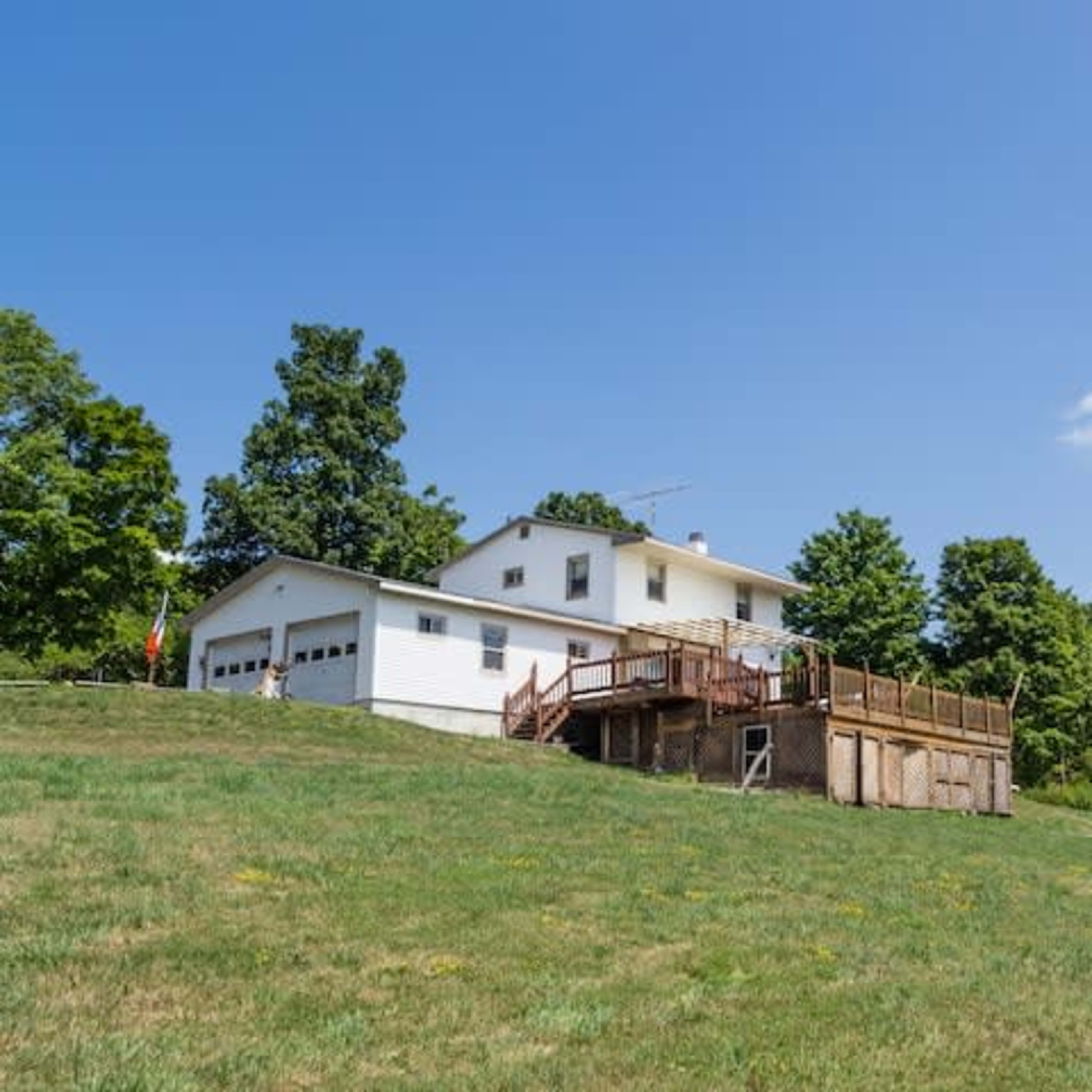 A two-story white house with a wooden deck and garage sits on a grassy hillside surrounded by trees.