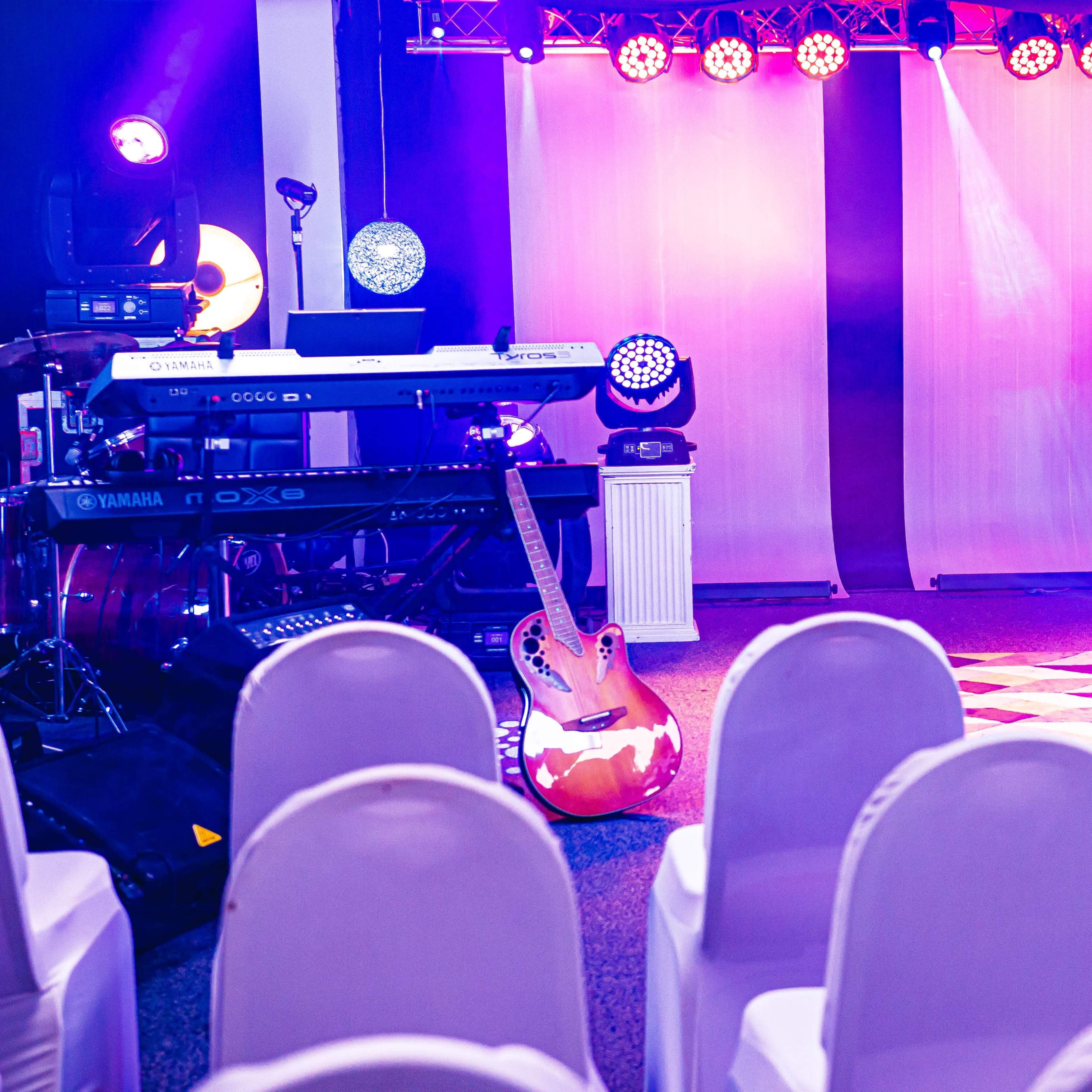 The scene features a stage setup with musical instruments, including a guitar and keyboard, in front of empty white chairs arranged for an audience.