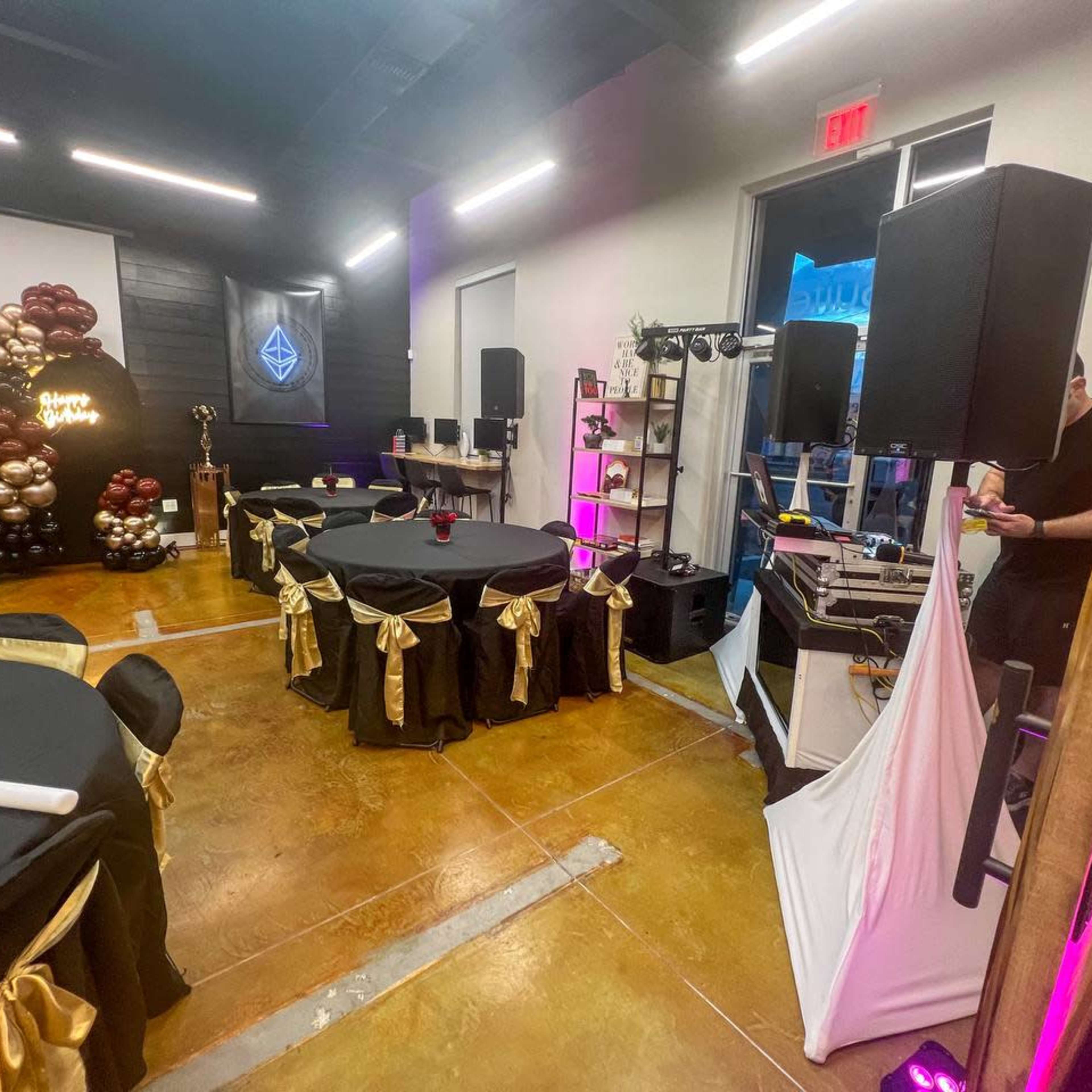 A party setup with black tables adorned with gold ribbons, a DJ booth, and decorative balloons in the background.
