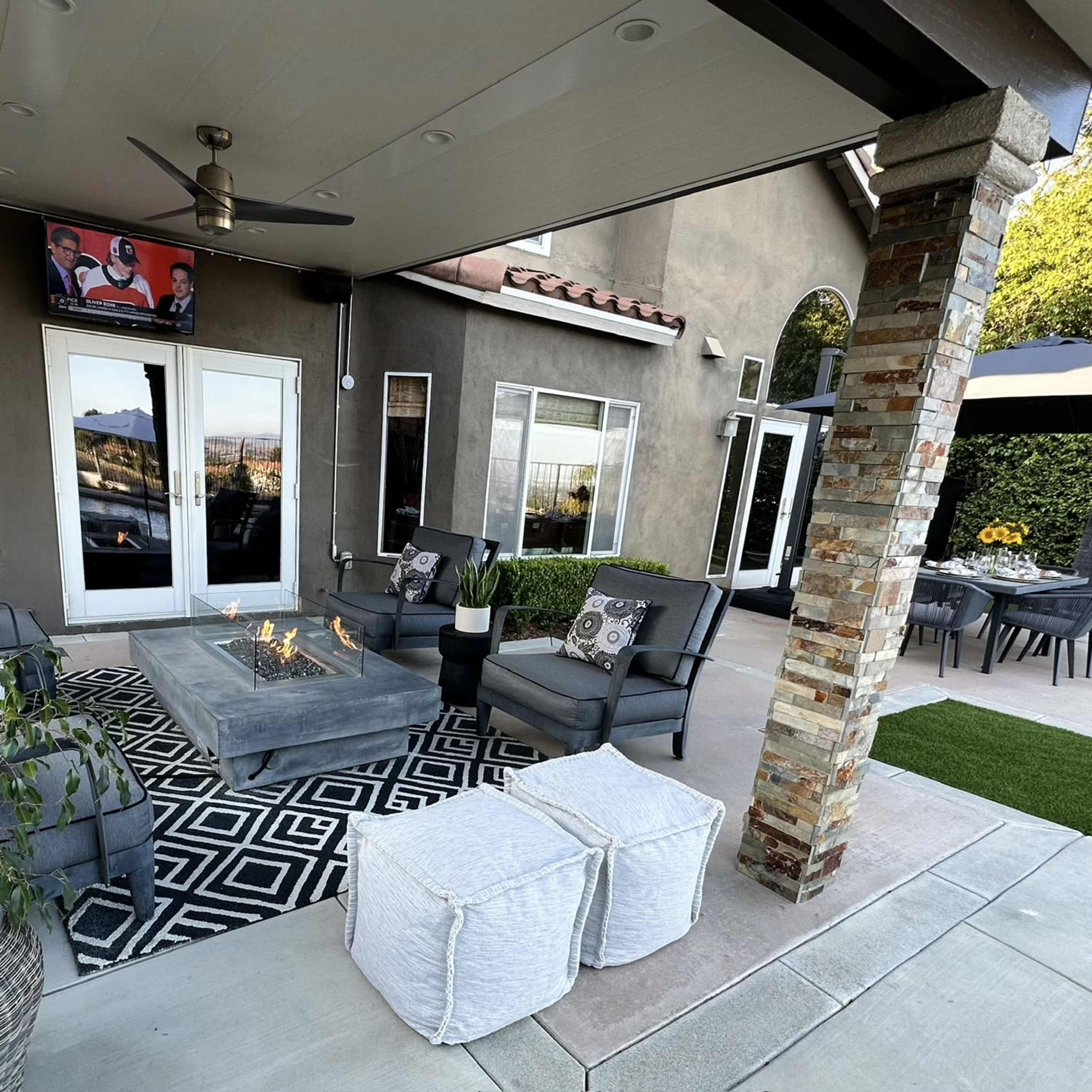 A patio area features a fire pit surrounded by seating, outdoor dining space, and a large umbrella, set against a landscaped backdrop.