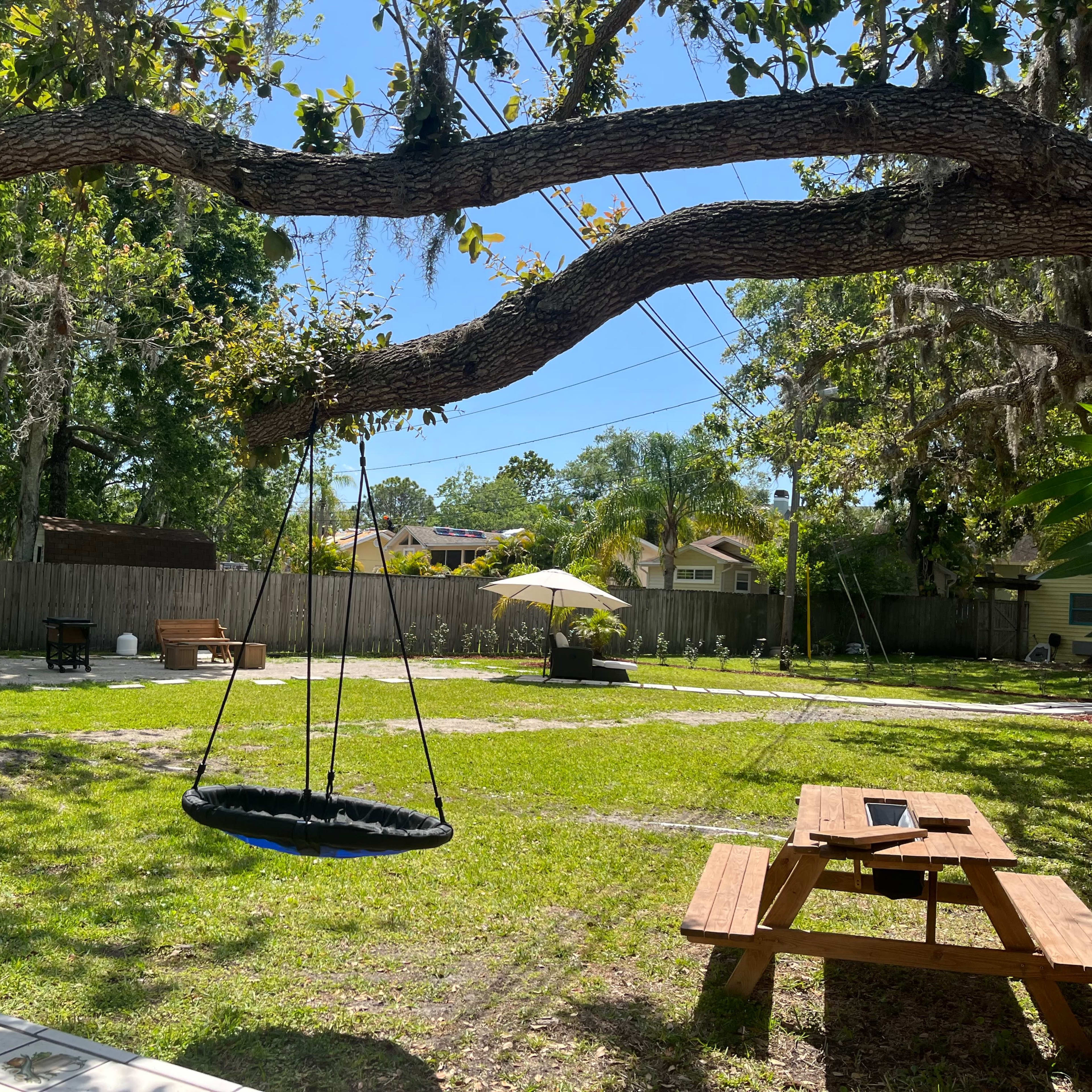 A swing hangs from a tree branch in a grassy backyard with picnic tables and an umbrella in the distance.
