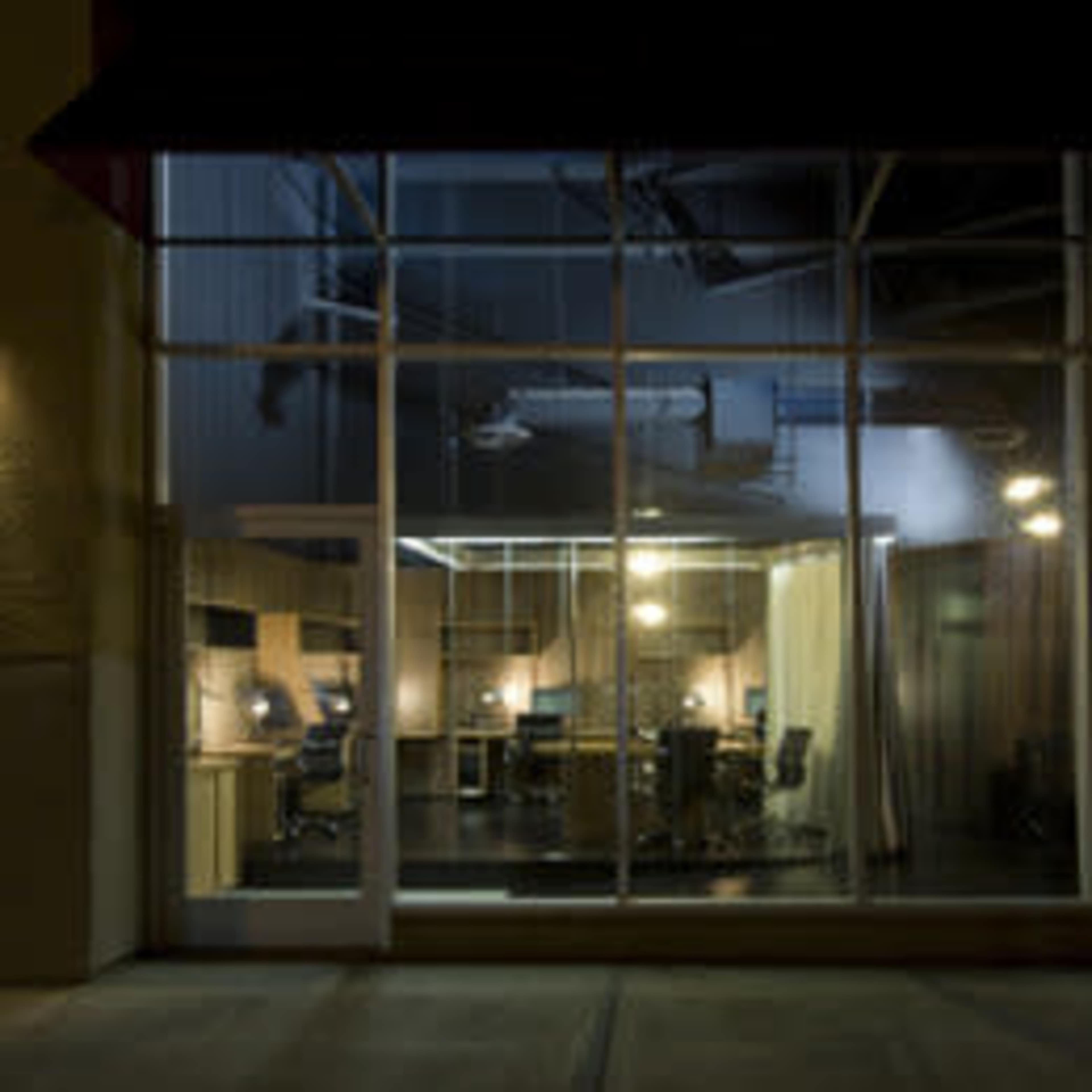 A dimly lit office space visible through large glass windows, with multiple workstations and chairs arranged inside.