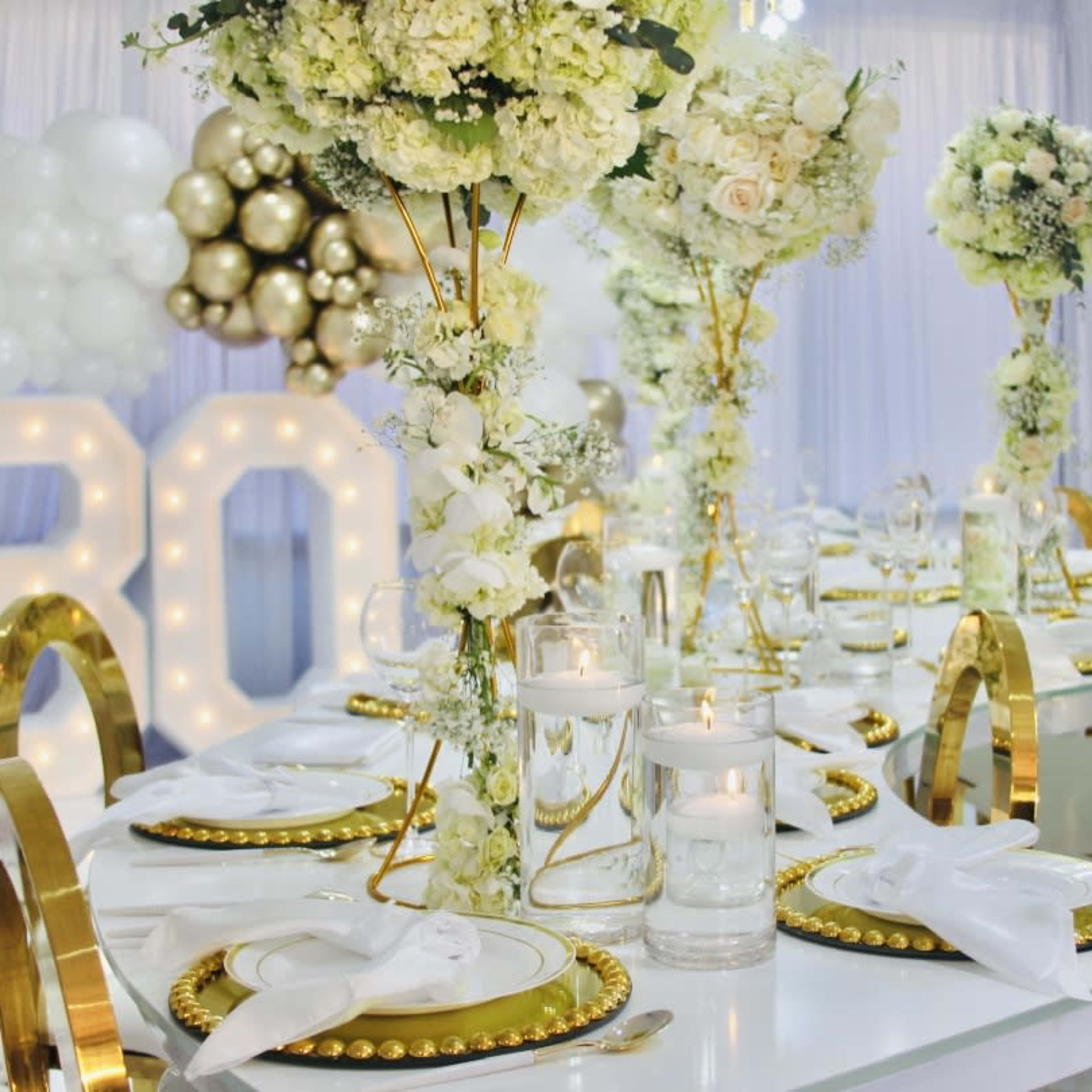 A elegantly arranged banquet table set for a celebration, featuring white floral centerpieces, gold accents, and large illuminated "80" in the background.