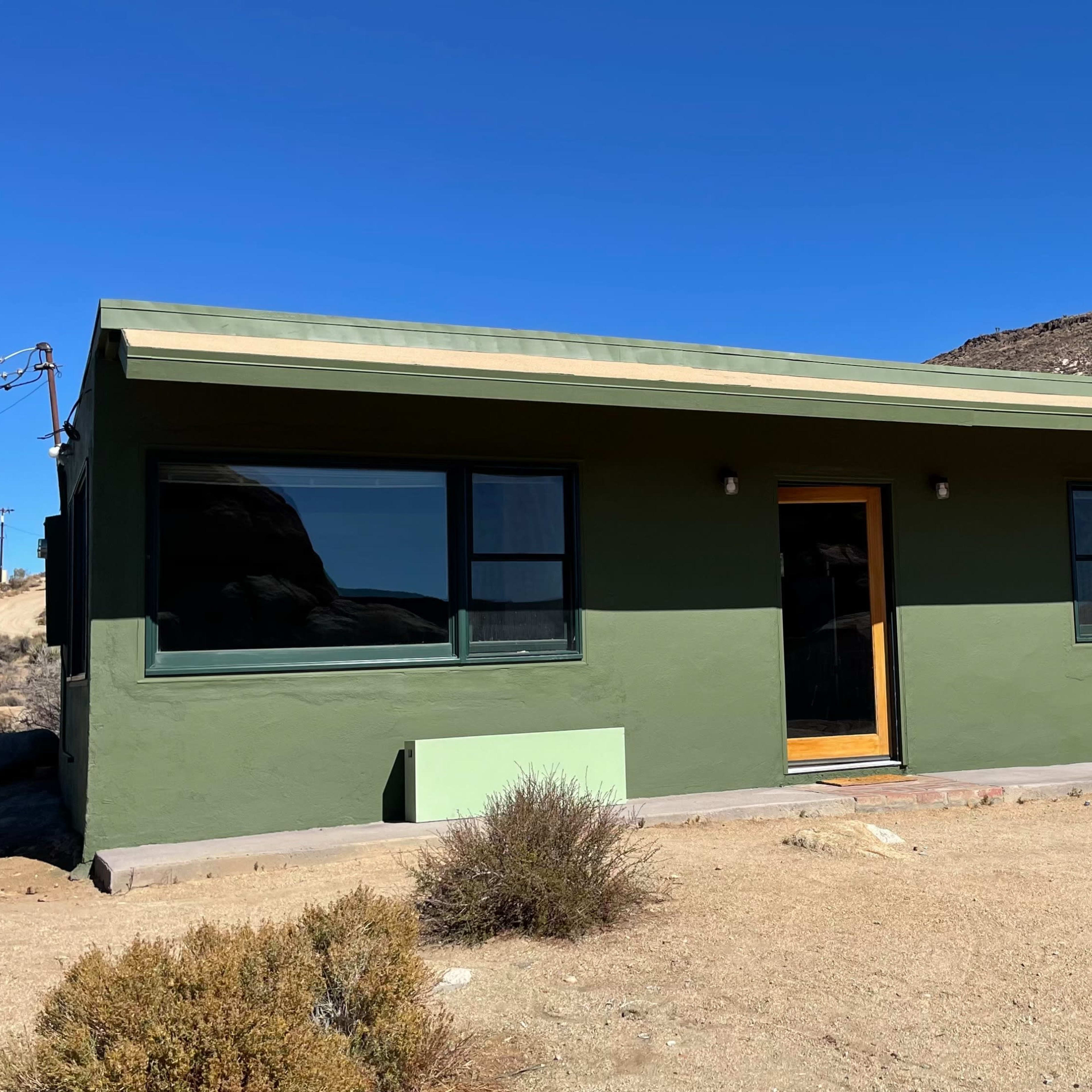 A single-story green house with large windows is situated on a sandy lot near a rocky hillside.