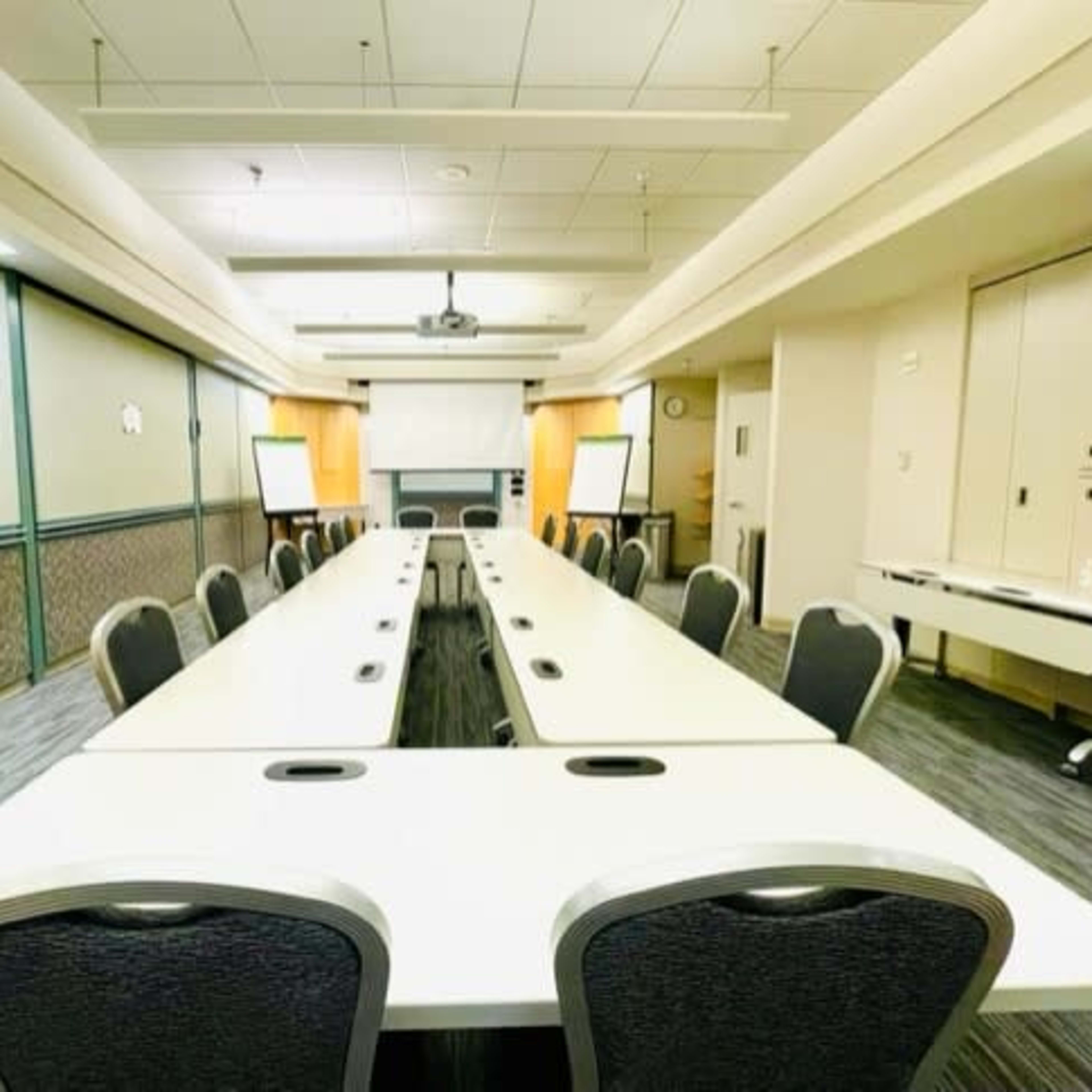 A large conference room features long tables arranged in a U-shape with chairs and presentation equipment at one end.