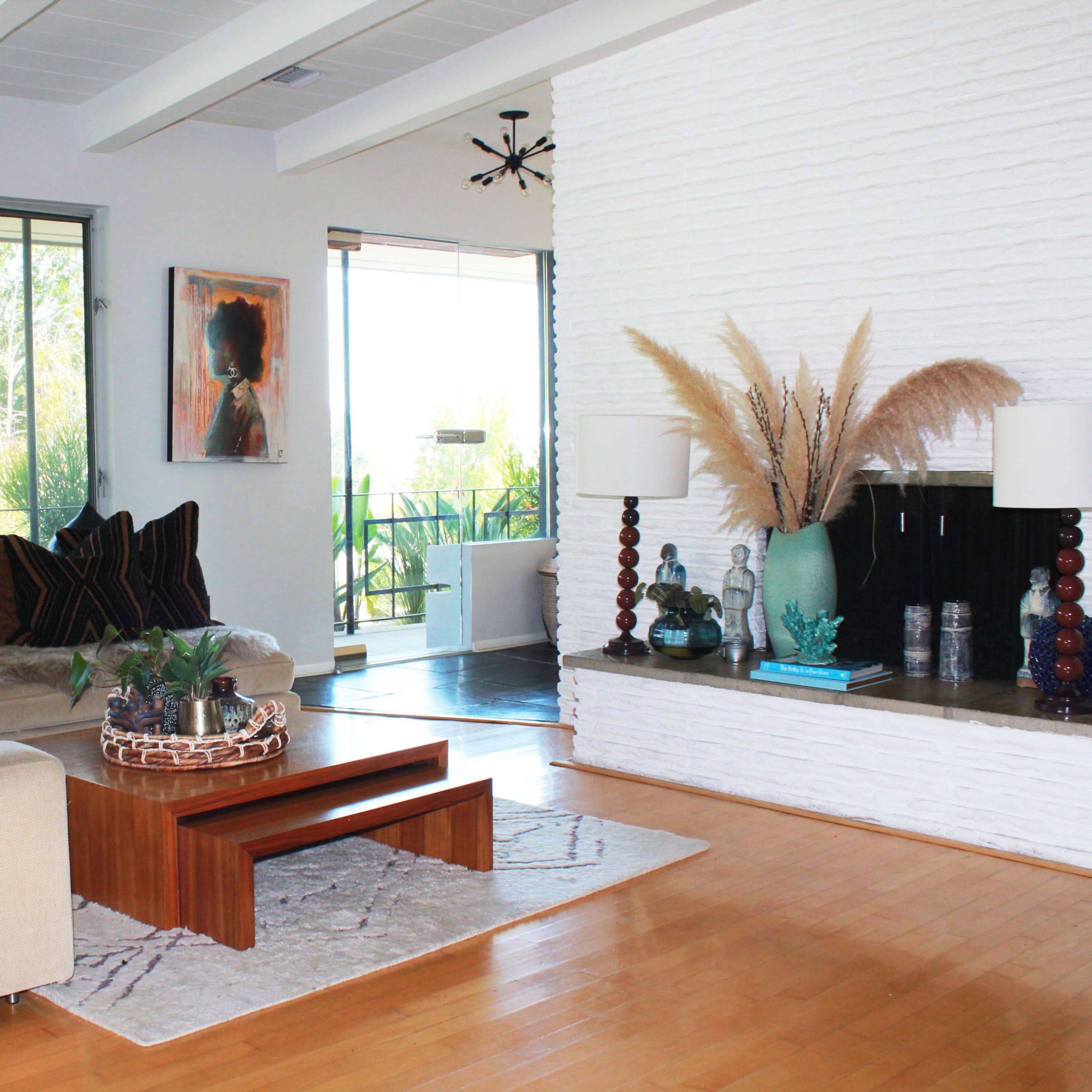 A modern living room features a beige sectional sofa with colorful pillows, a wooden coffee table, and a decorative area rug, while large windows provide views of greenery.