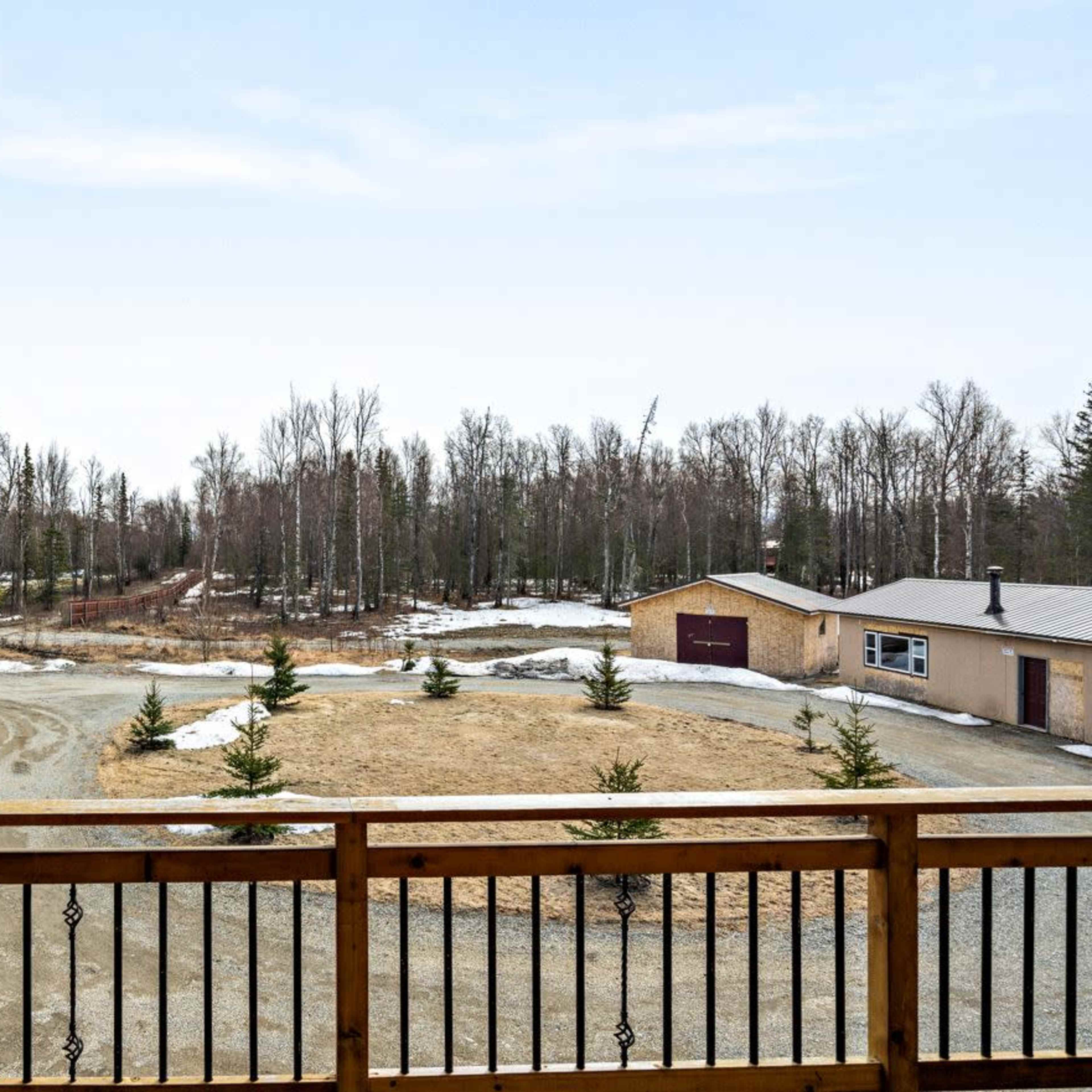 A rural landscape with a gravel driveway, a house with a garage, and sparse trees in the background, surrounded by patches of snow.