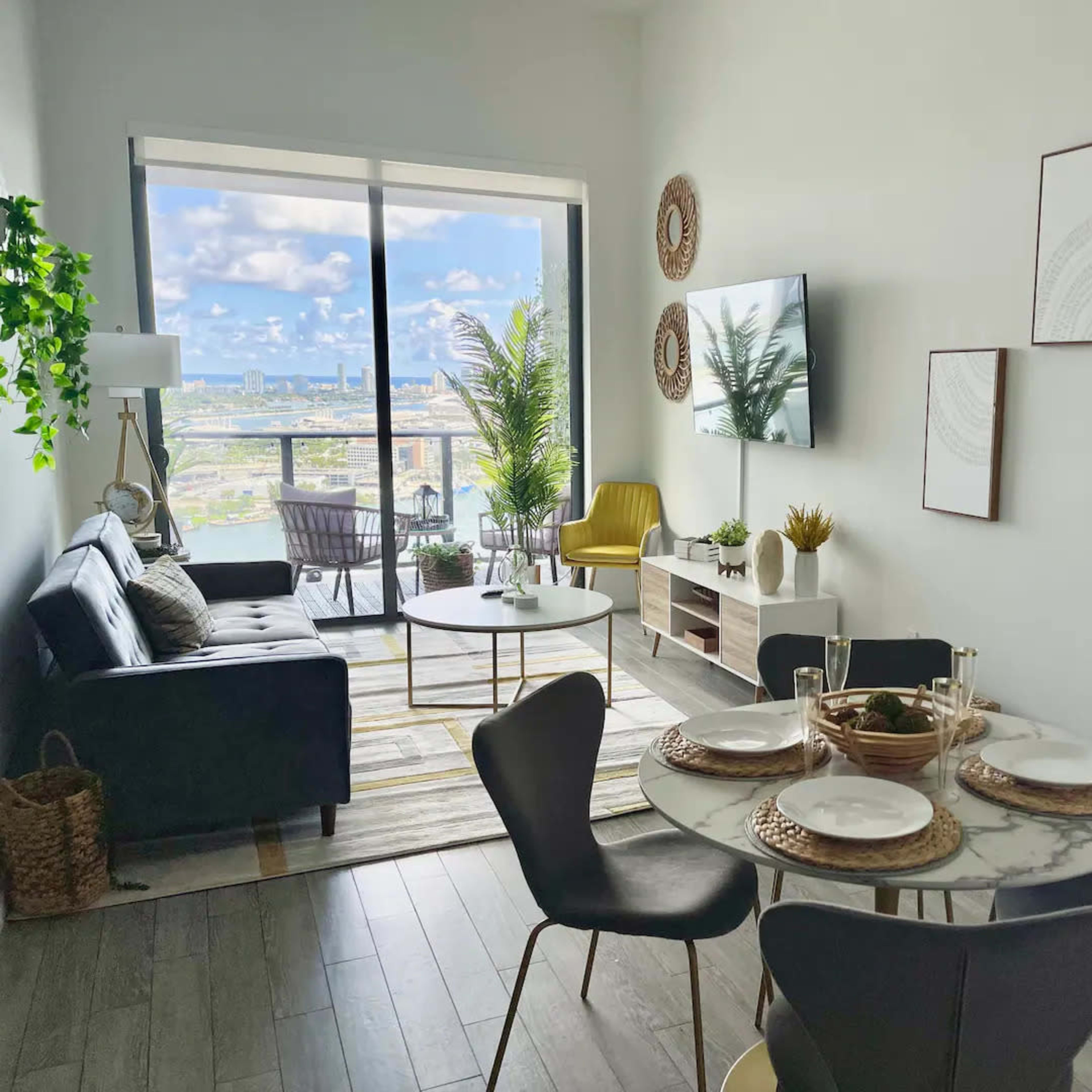 A modern living room with a dining area features a black sofa, round marble table, and large windows providing a view of the cityscape.