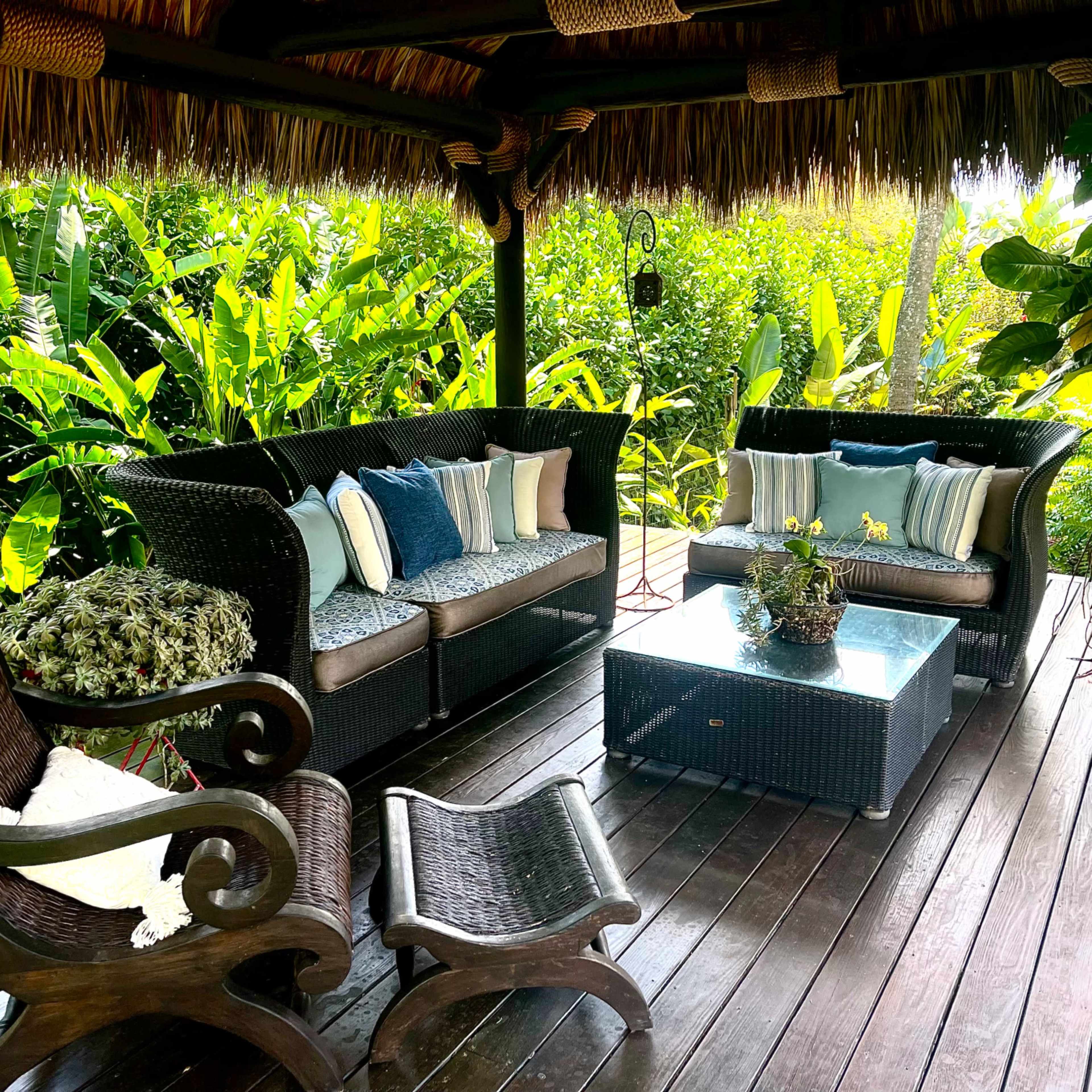A covered outdoor seating area features wicker furniture, a coffee table, and lush greenery in the background.
