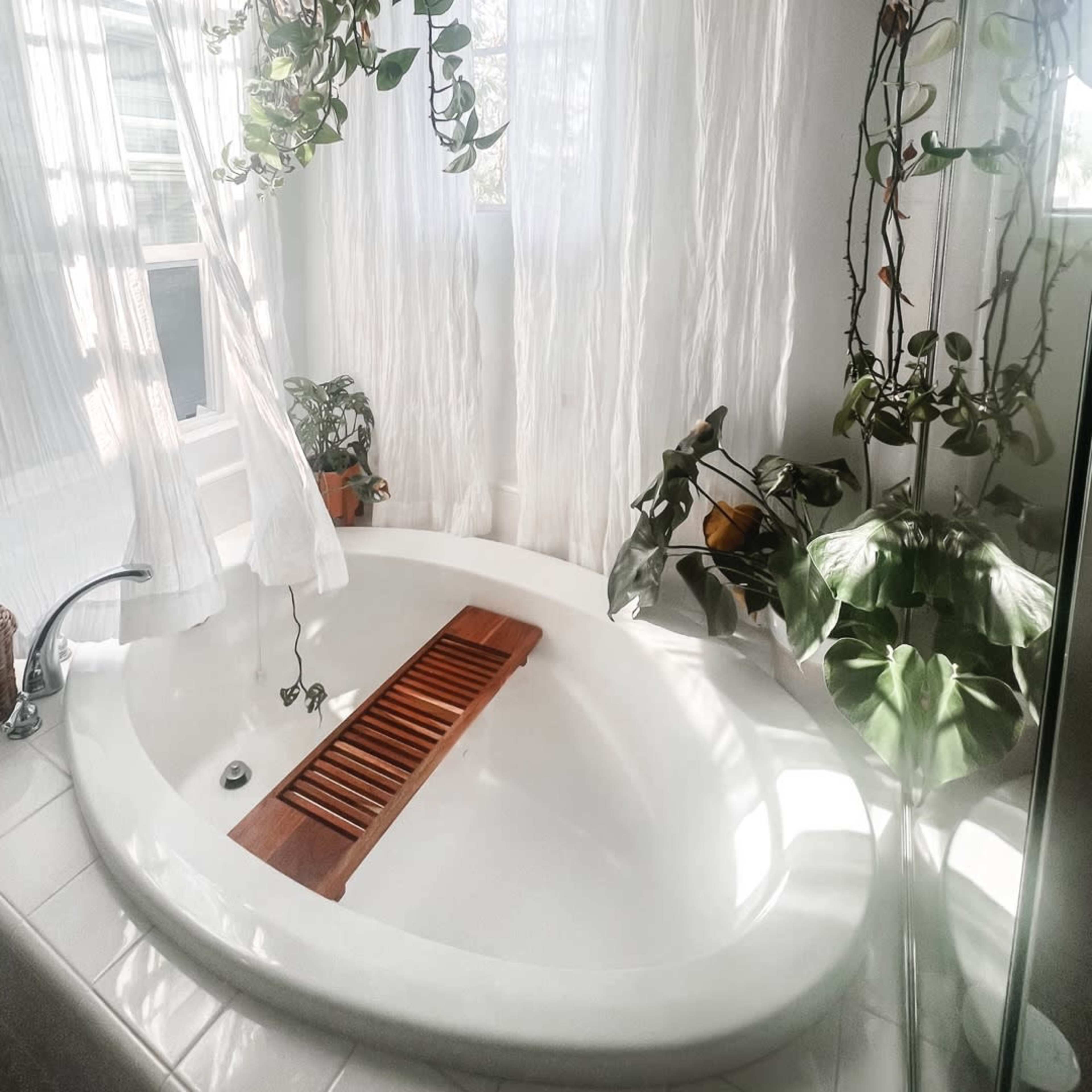 A curved white bathtub is surrounded by plants and sheer white curtains, with a wooden slatted cover resting on top.