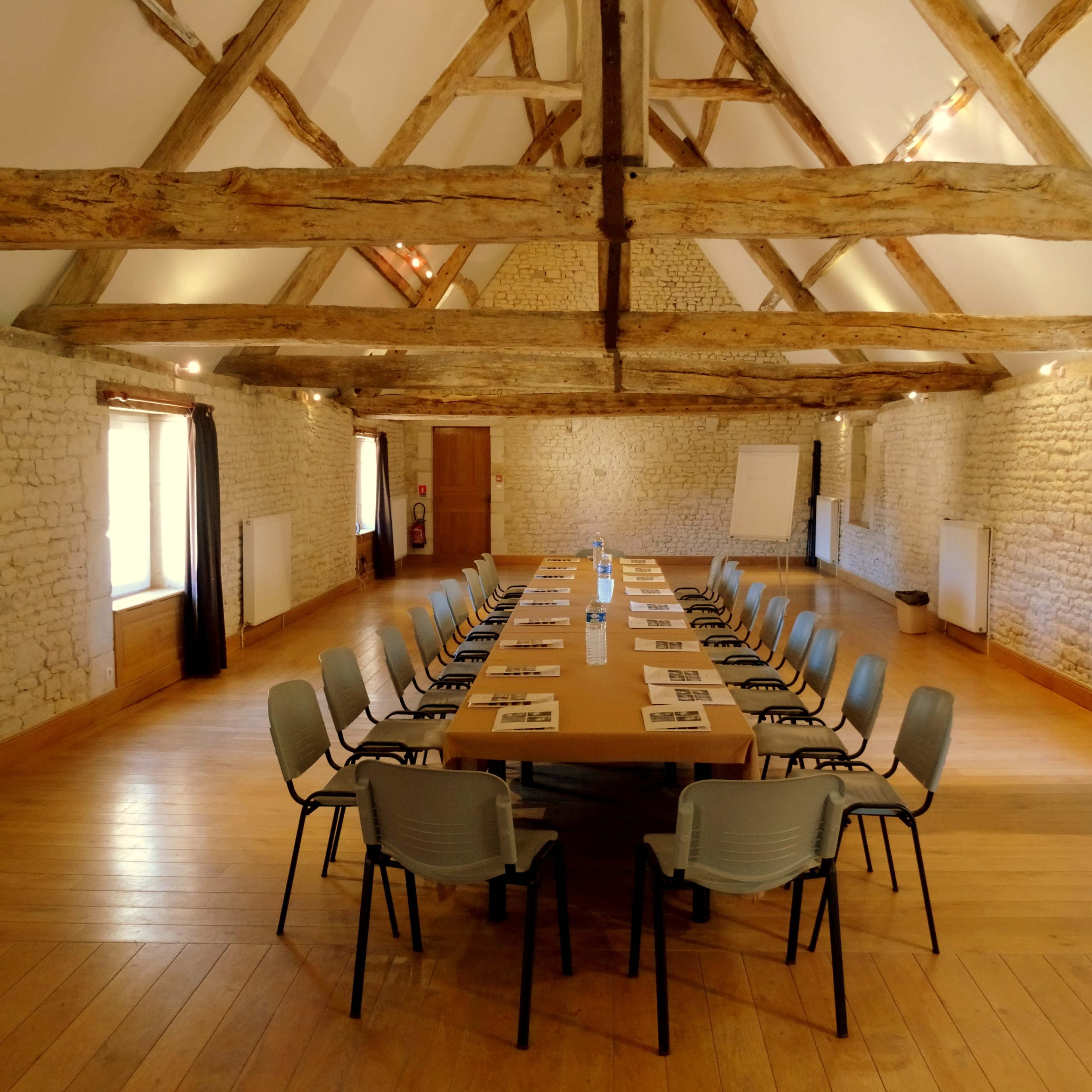 A long wooden table is surrounded by gray chairs in a spacious room with exposed wooden beams and stone walls.