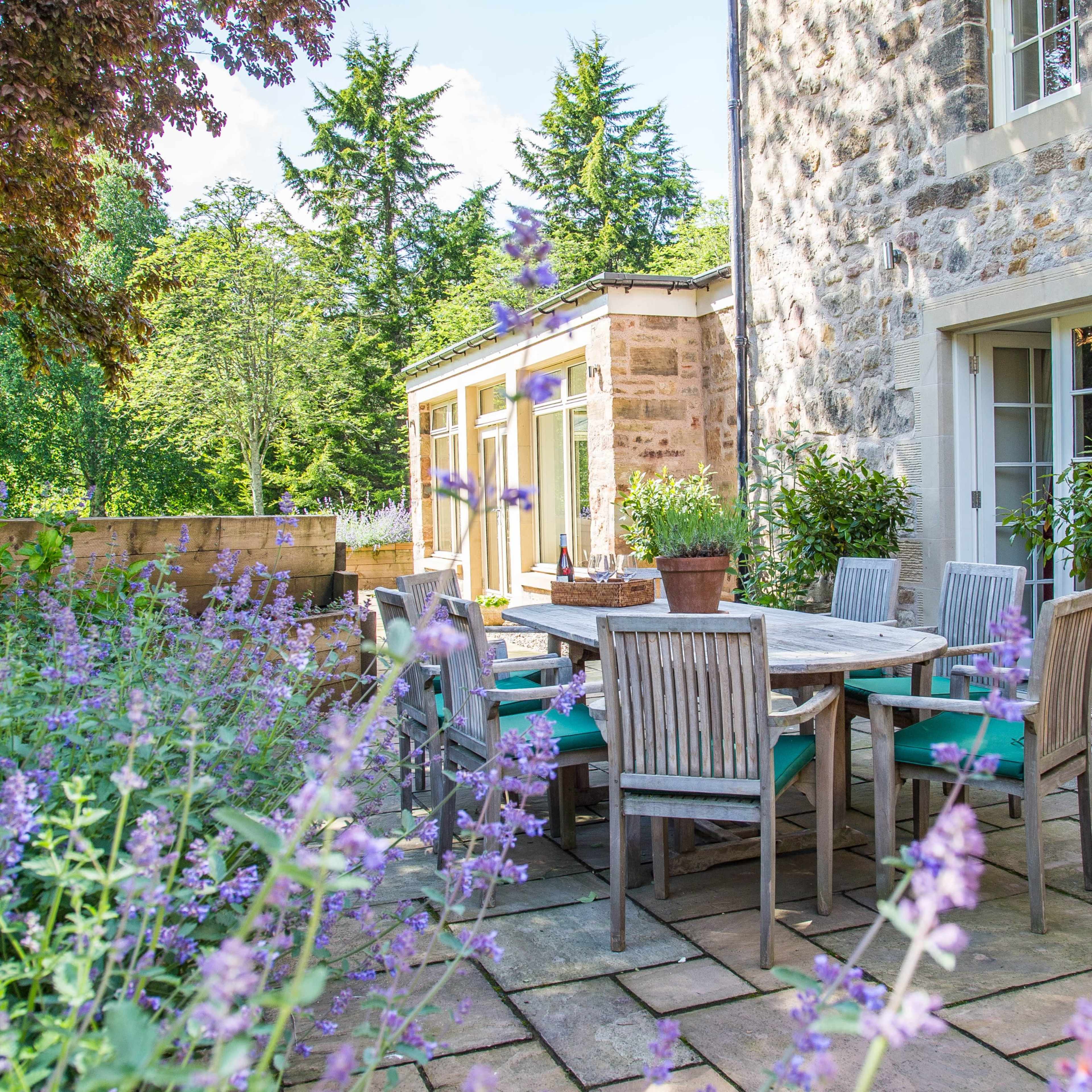 A stone house features a patio with a wooden dining set surrounded by potted plants and a garden with blooming flowers.