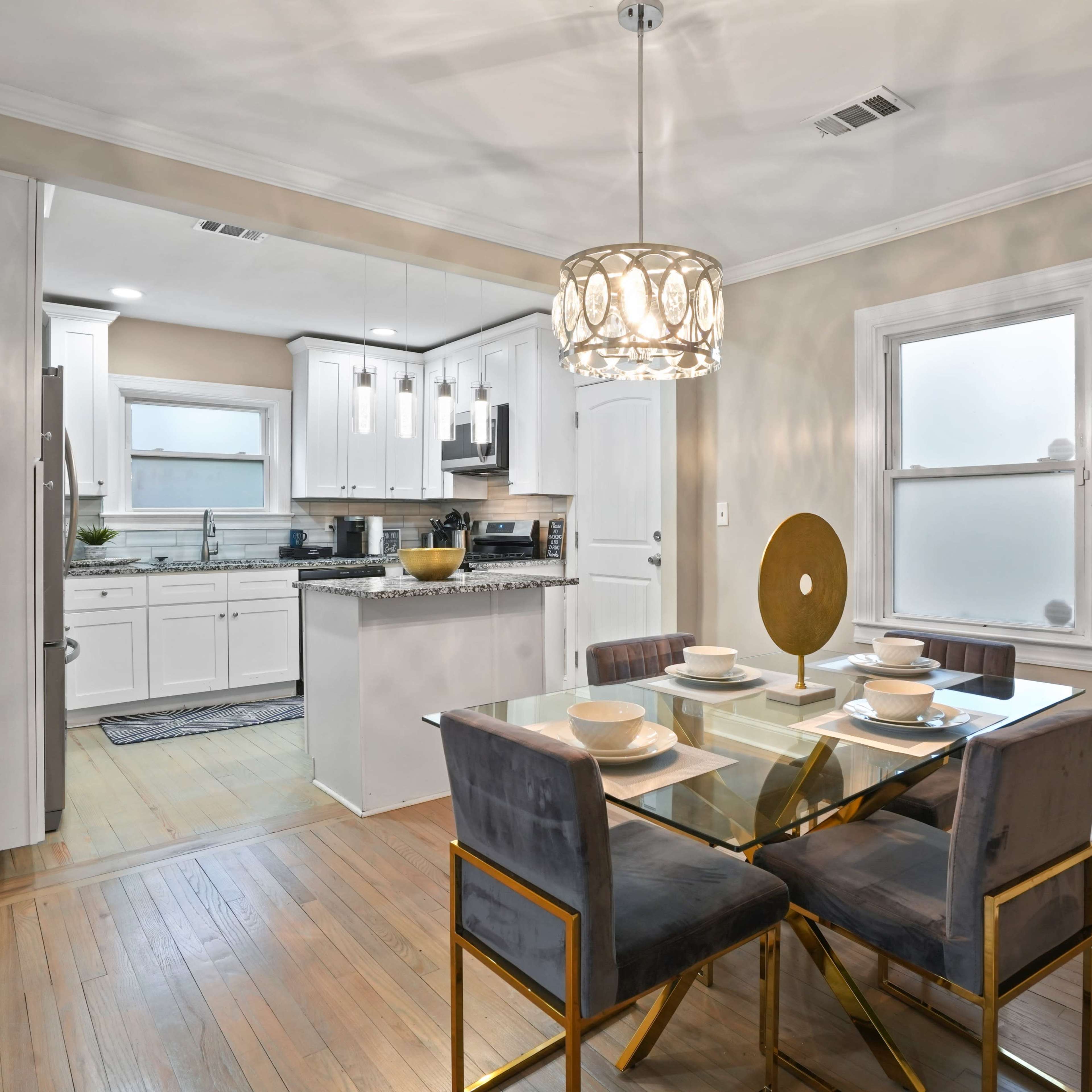 A modern dining area features a glass table set for four with metallic chairs, leading to a kitchen with white cabinetry and granite countertops.