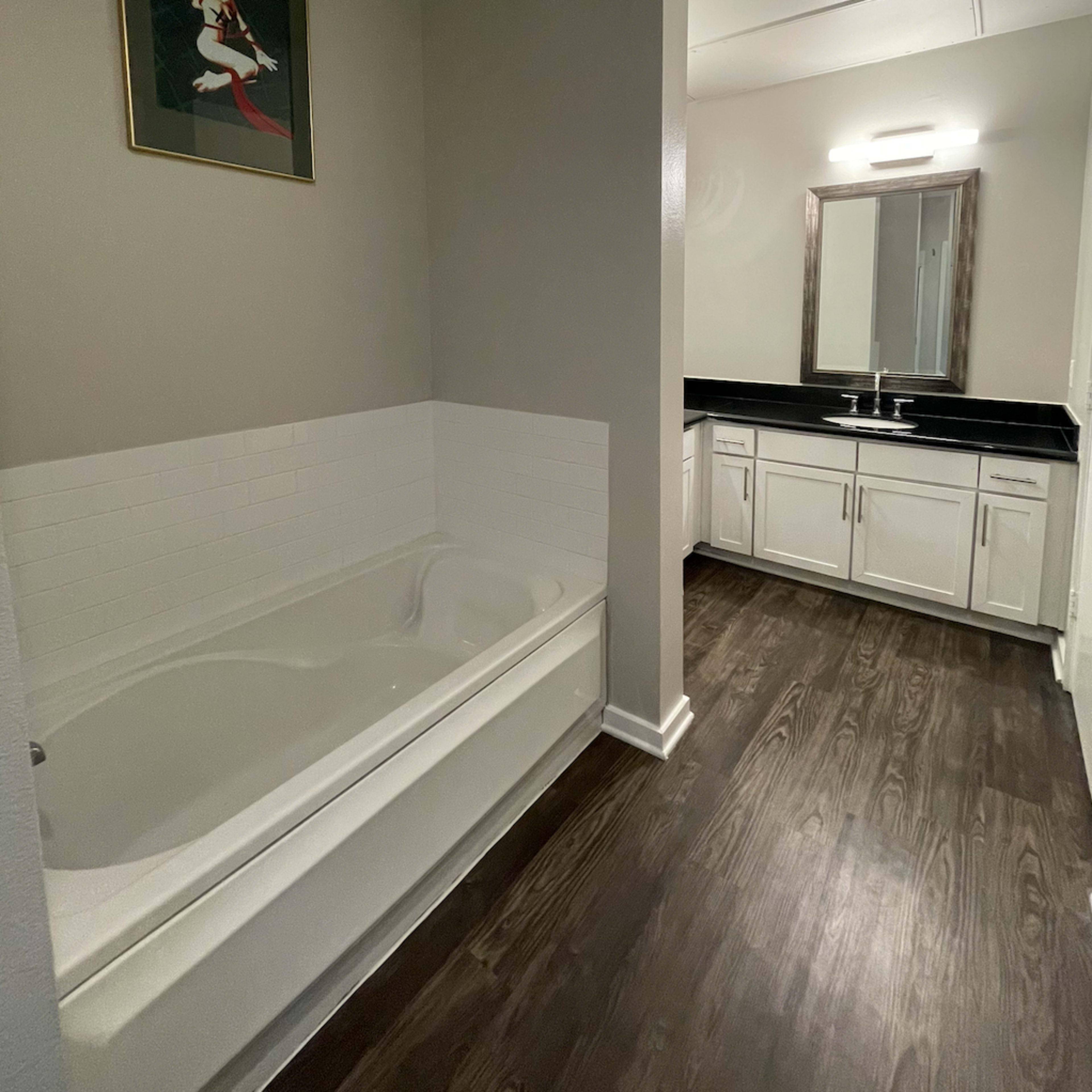 A bathroom features a bathtub on the left and a double vanity with a mirror on the right, all set against a neutral-colored wall and dark wood flooring.