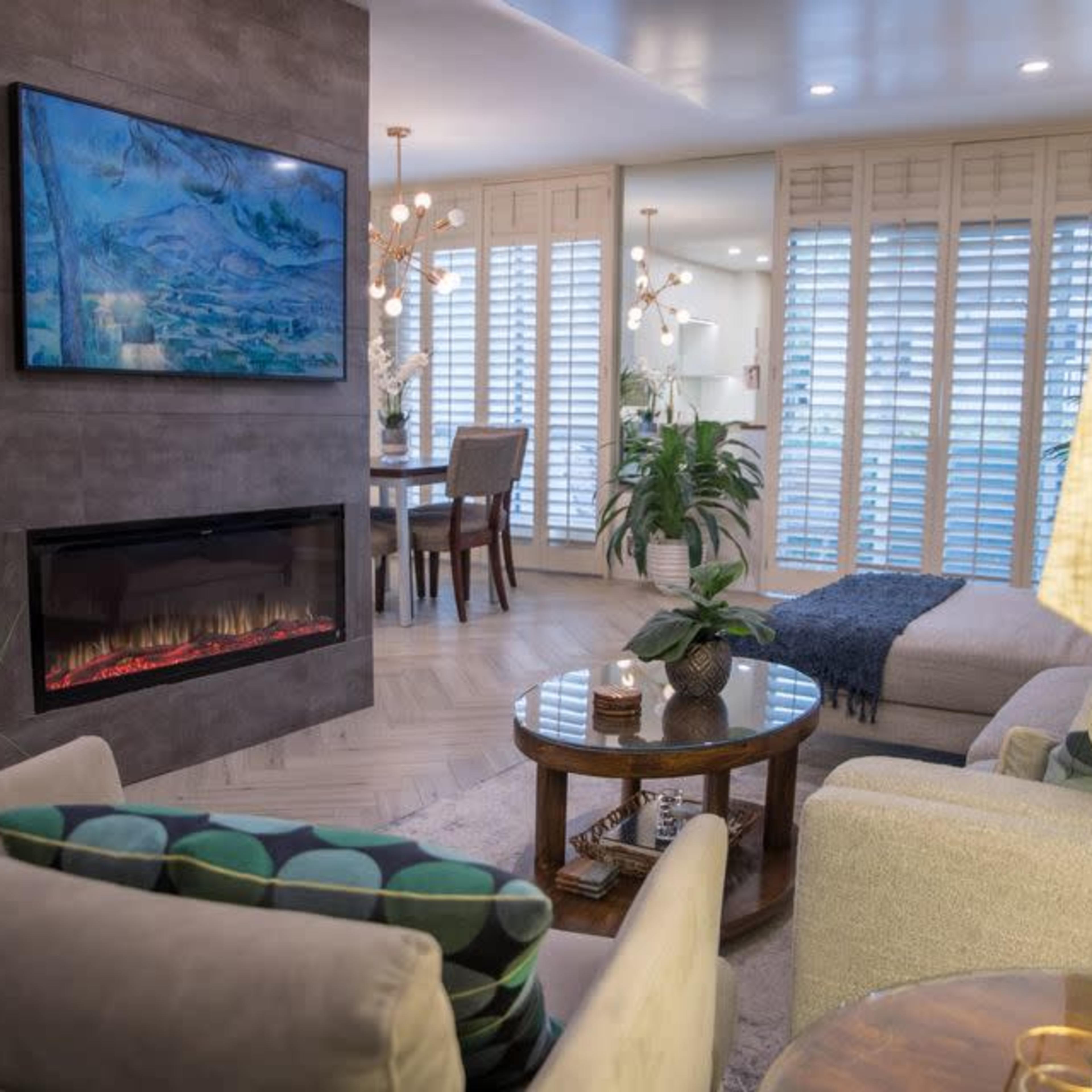 A modern living room featuring a television over a fireplace, wood flooring, and a dining area in the background.