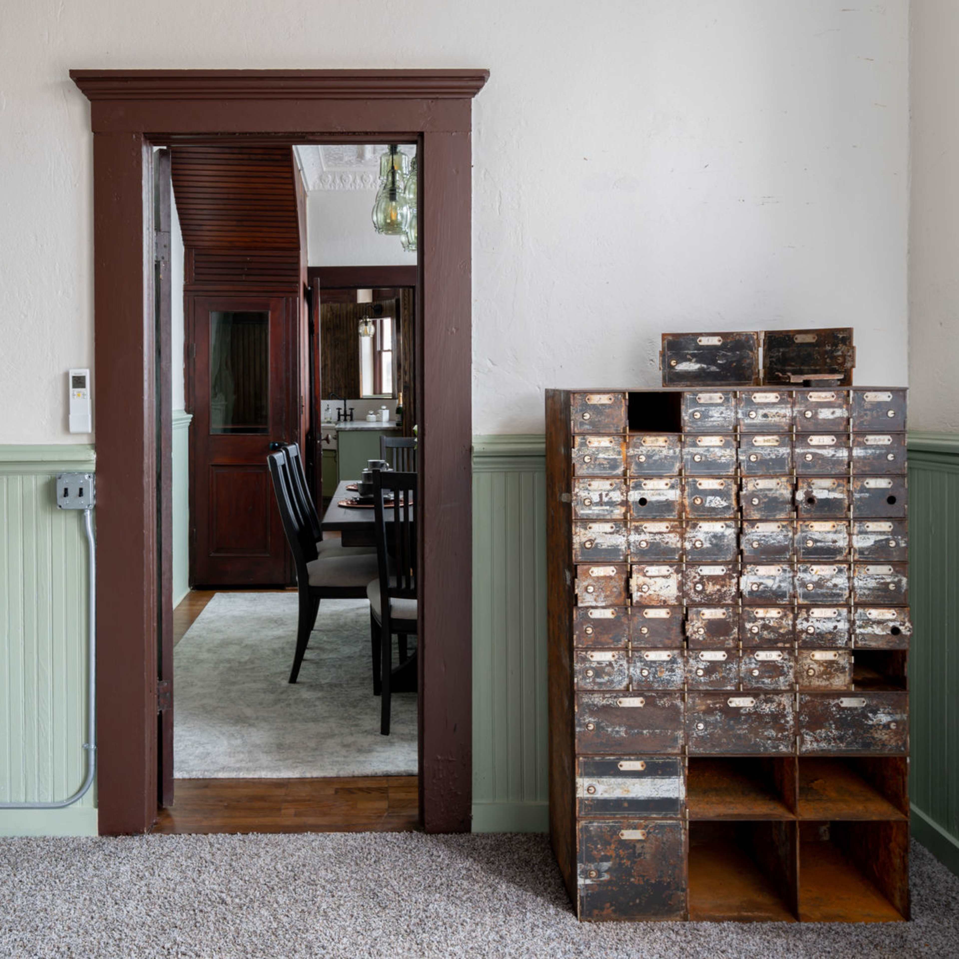 A room with a door leading to another space, framed by dark wooden trim, alongside a multi-drawer wooden cabinet against a green wall.
