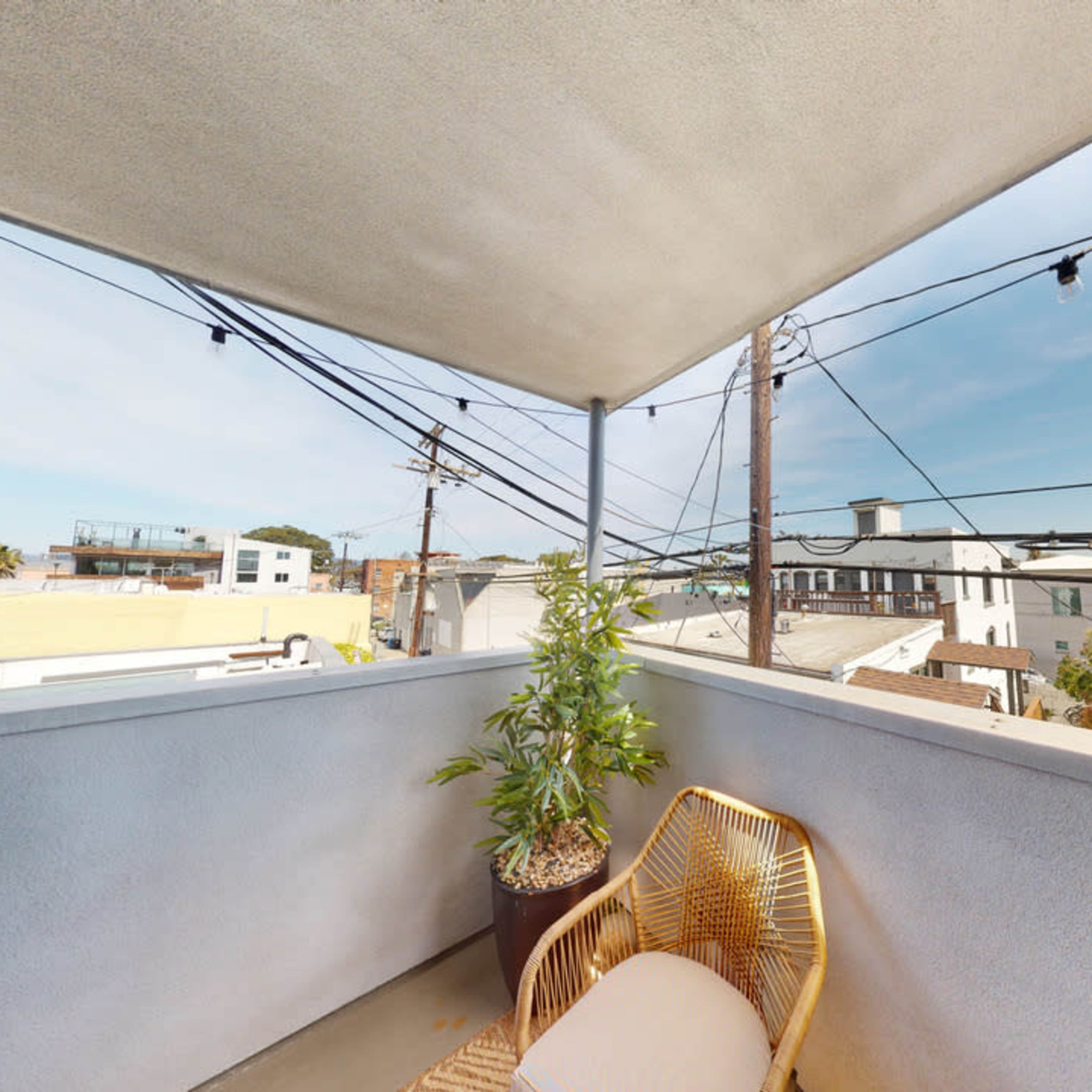 A small balcony featuring a potted plant and a chair, with utility poles and buildings visible in the background under a clear sky.