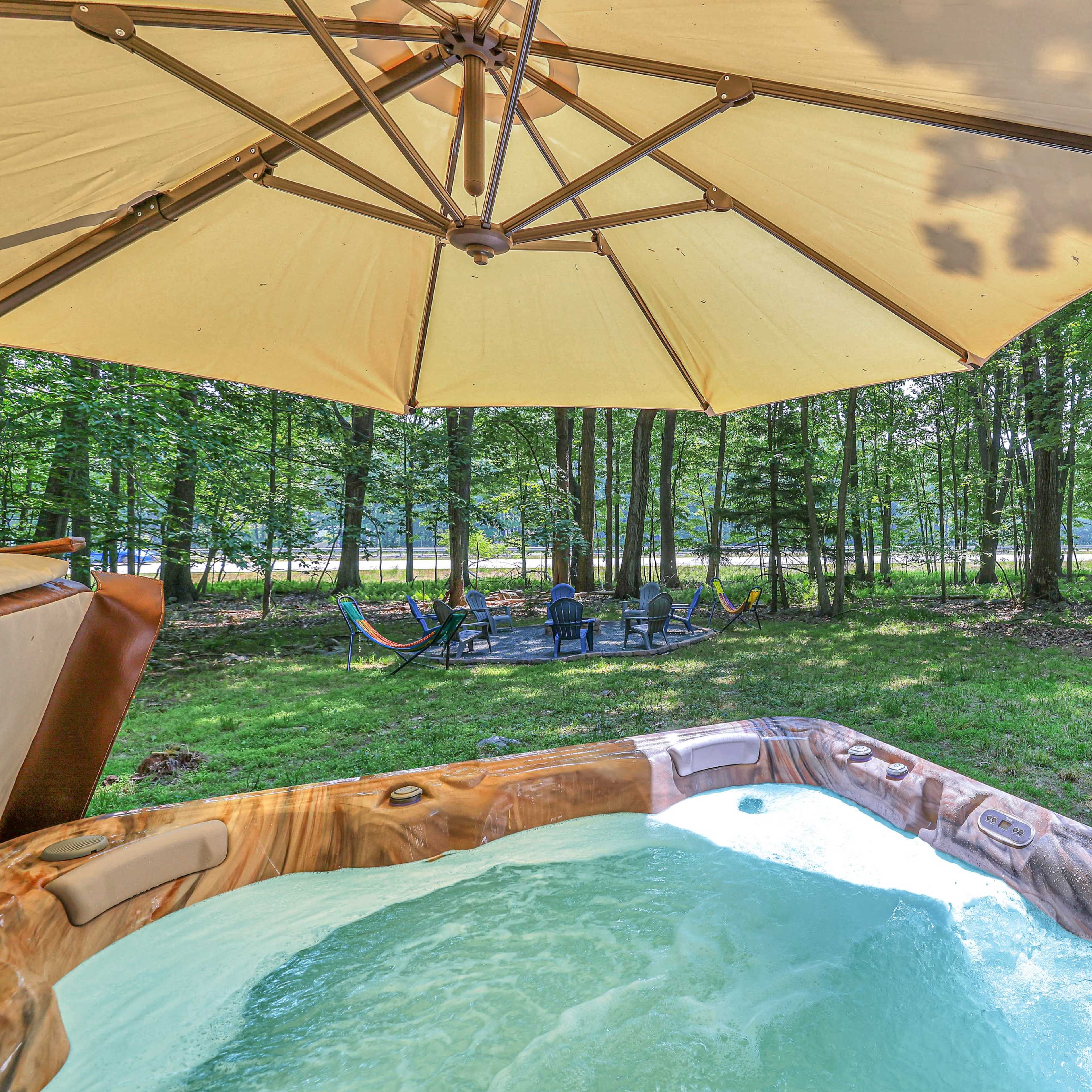 A hot tub is situated under a large umbrella, with a view of a forested area and a hammock nearby.
