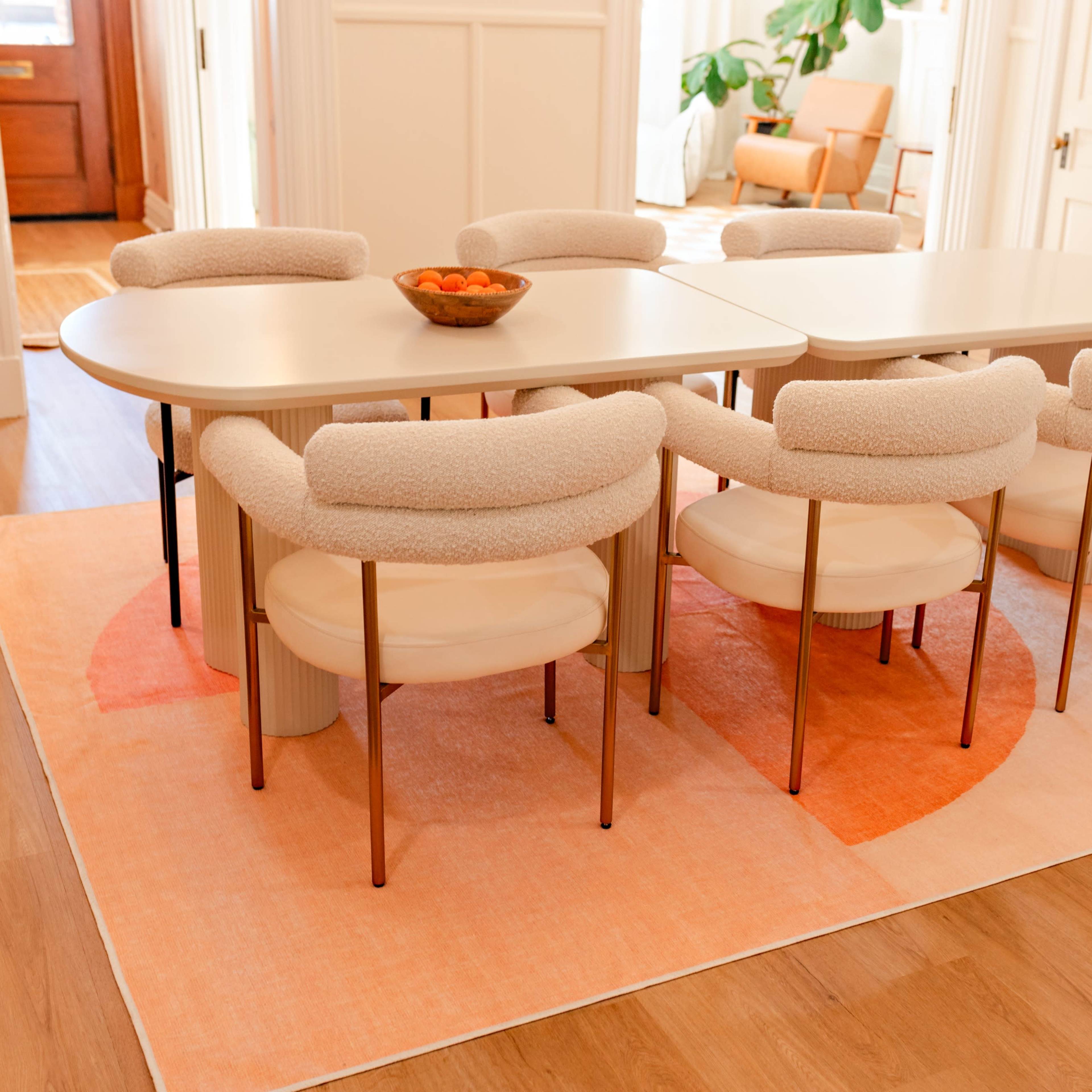 A modern dining table surrounded by upholstered chairs is set on a large, colorful area rug in a well-lit room.
