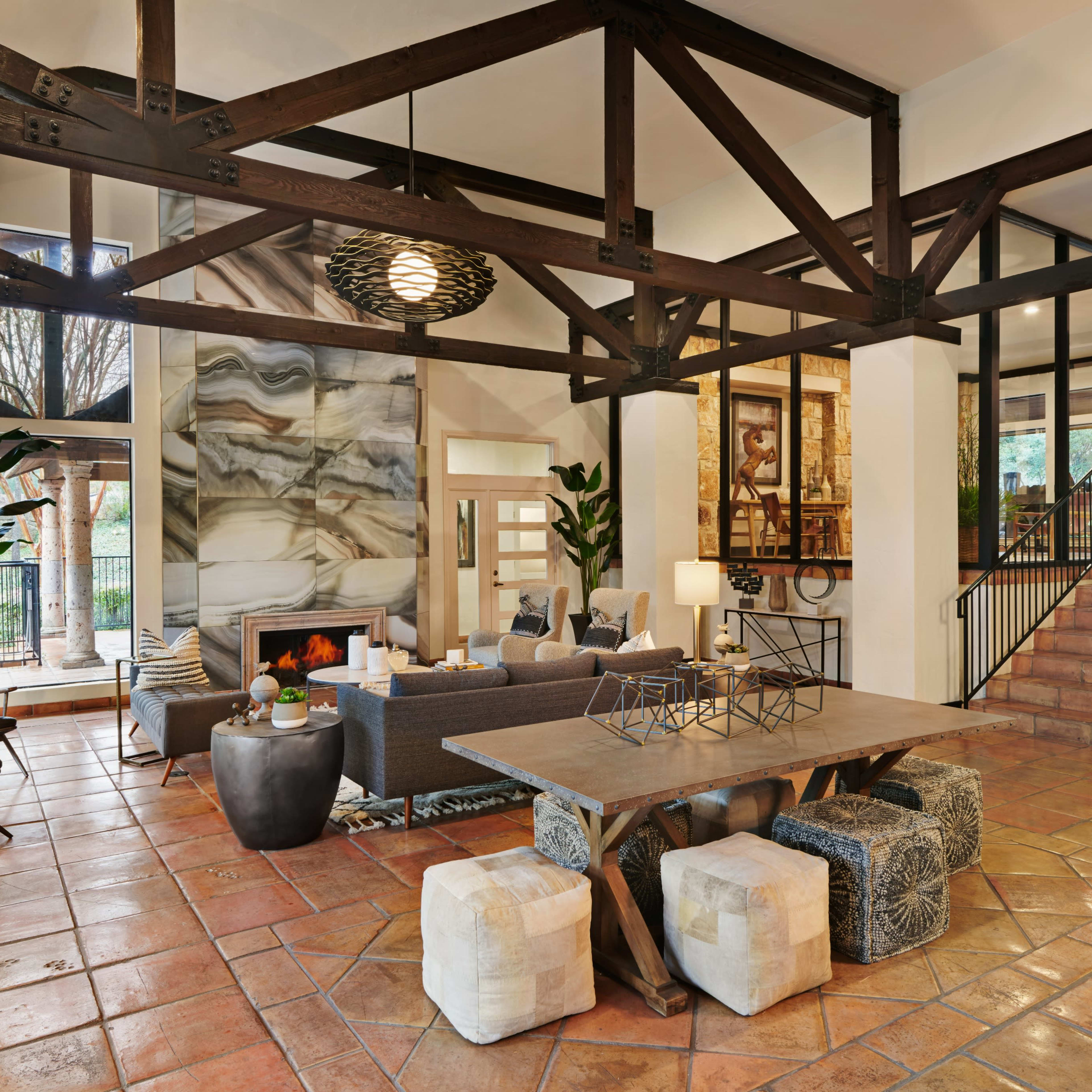 A spacious living area with a high ceiling, featuring large windows, a stone fireplace, and a mix of modern furniture, including a large table and comfortable seating.