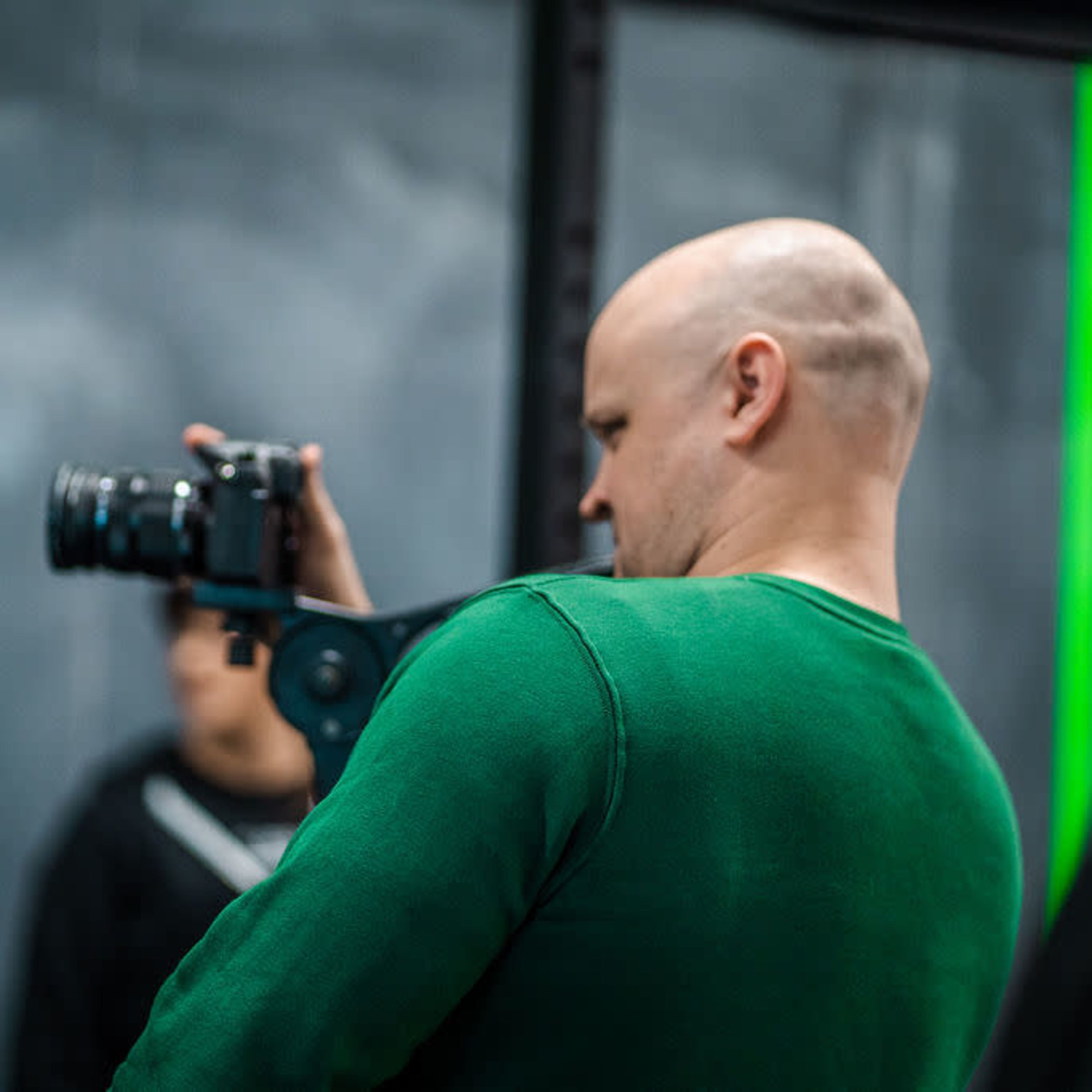A man in a green shirt holds a camera while facing a dark background with a green line, and another person is partially visible behind him.
