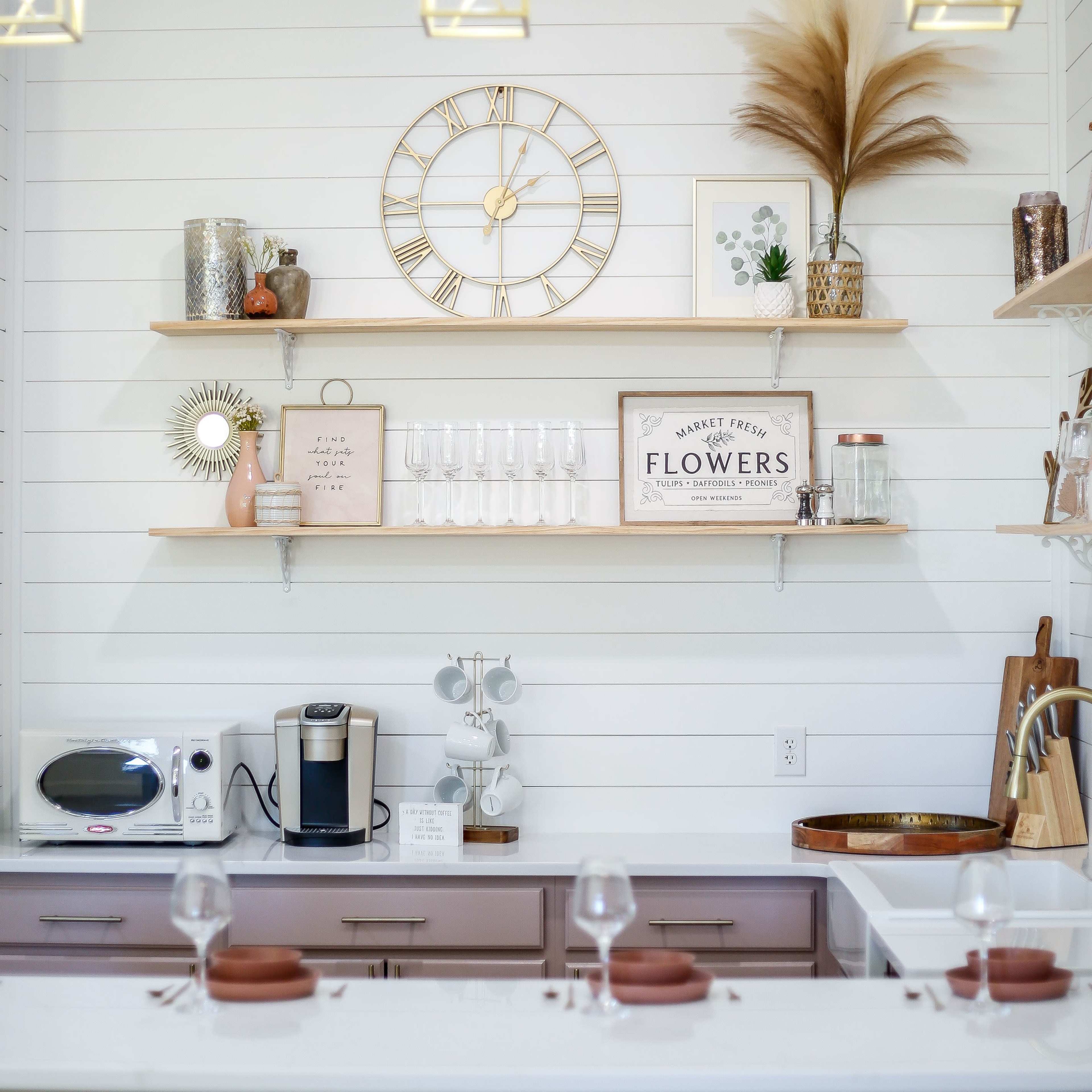 A modern kitchen scene features wooden shelves holding decorative items, a clock, and glassware, with a coffee maker on the countertop.