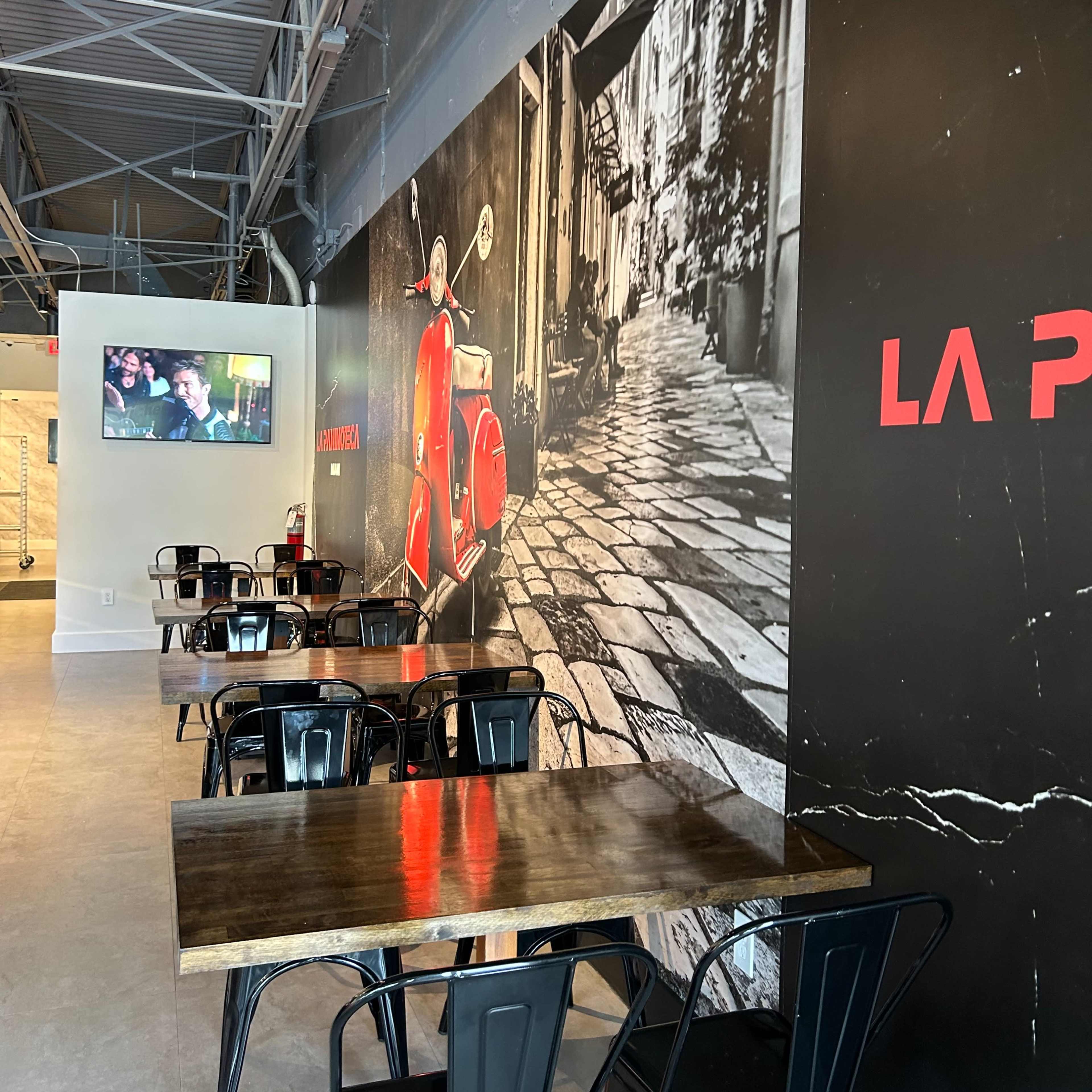 A modern cafe interior with black metal chairs and wooden tables, featuring a large mural of a red scooter on a cobblestone street.