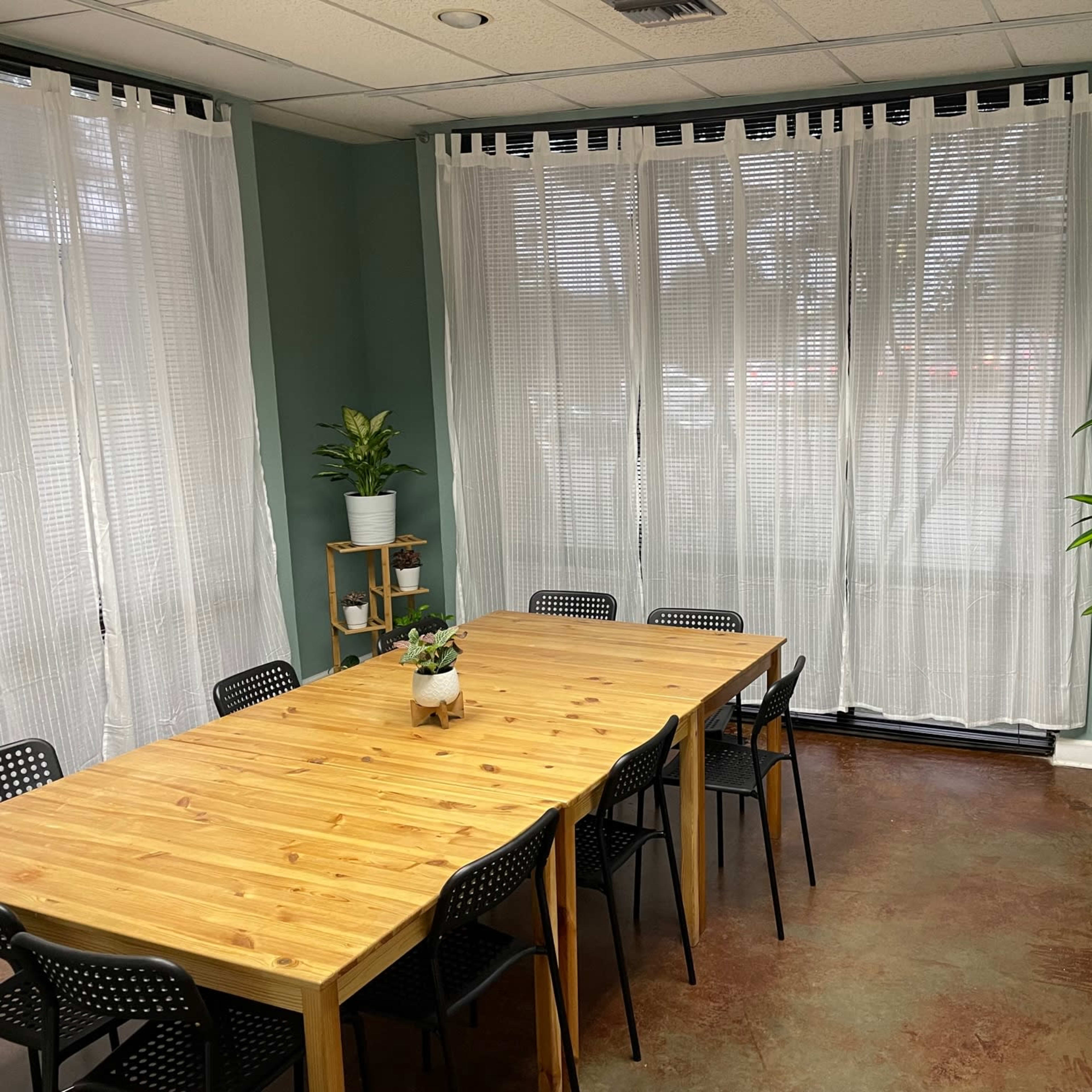 A conference room with a large wooden table surrounded by black chairs, brightened by natural light from large windows with sheer curtains.