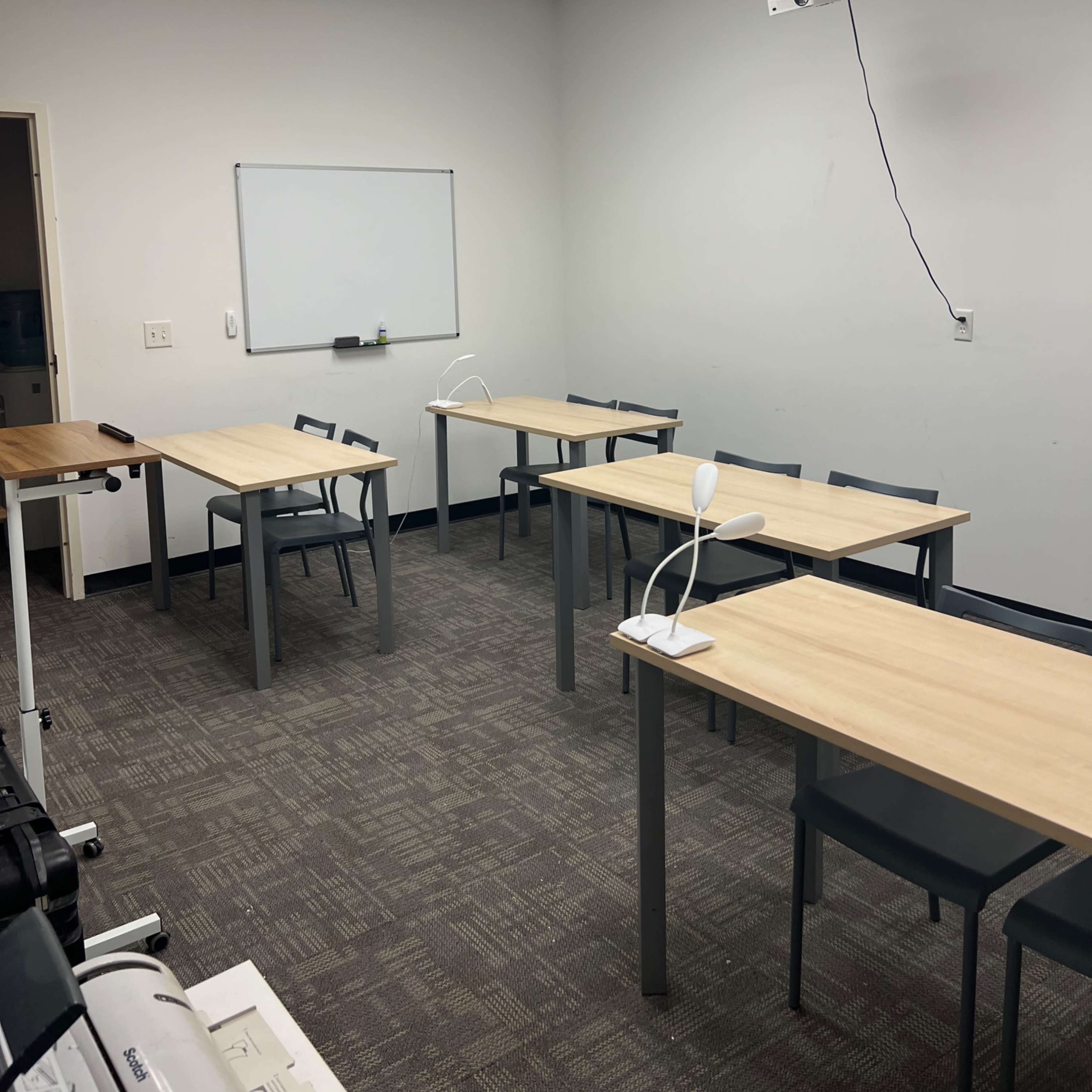 A classroom with four tables, each accompanied by a chair, a whiteboard on the wall, and a projector mounted from the ceiling.