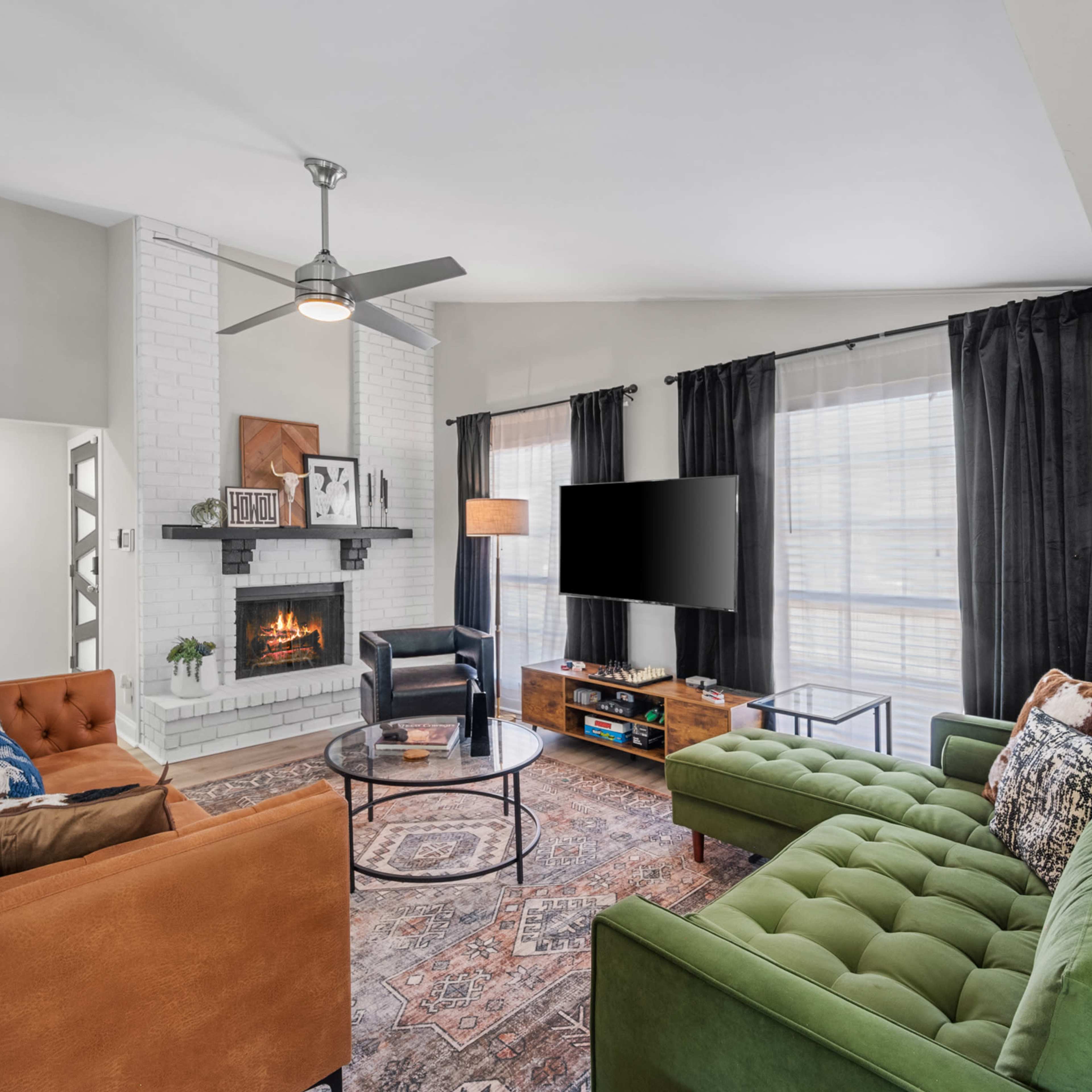 A living room features a cozy seating arrangement with an orange and green couch, a round coffee table, a white brick fireplace, and a television mounted on the wall.