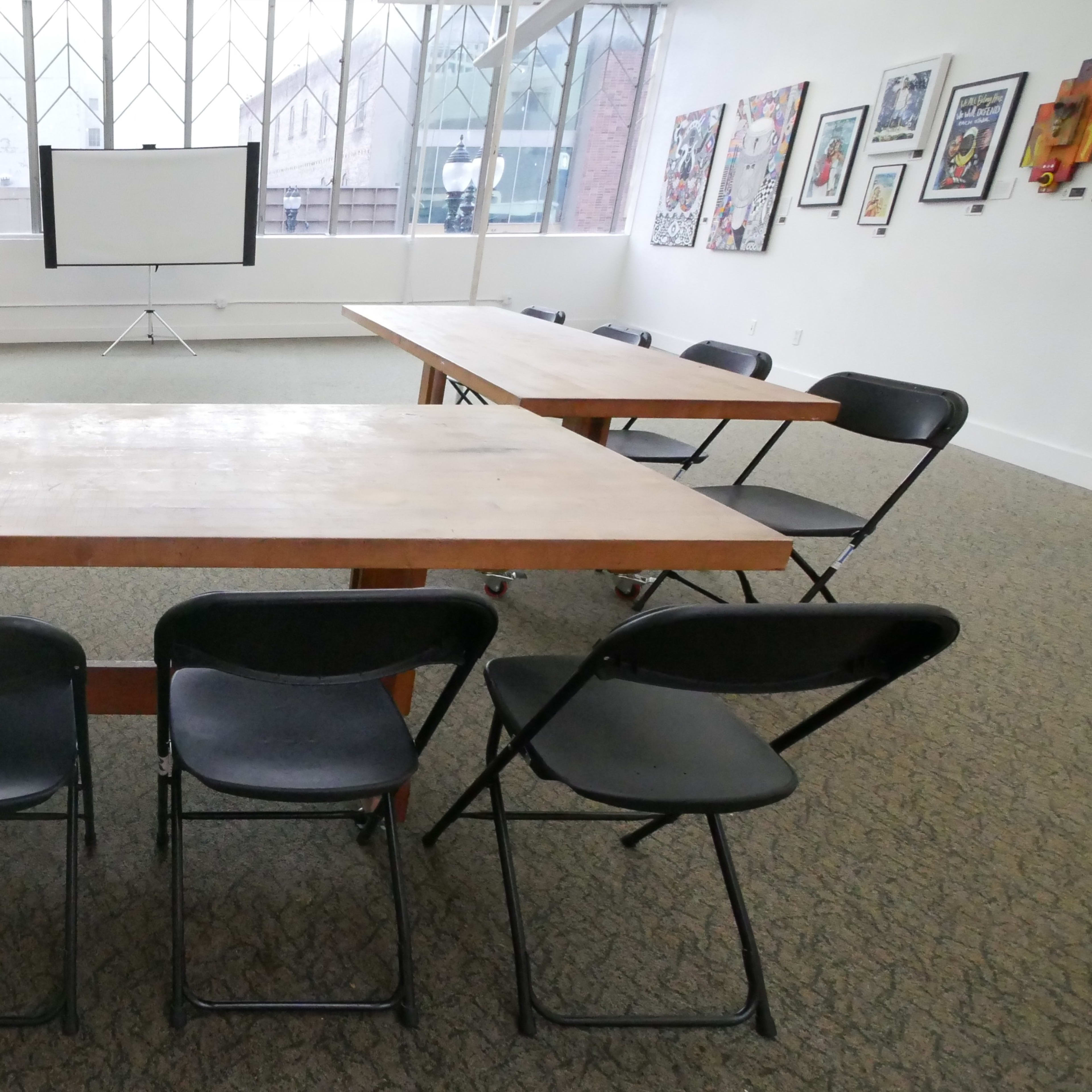 A spacious room features two large, rectangular tables arranged in a U-shape, surrounded by black folding chairs, with a projector screen positioned at the far end.