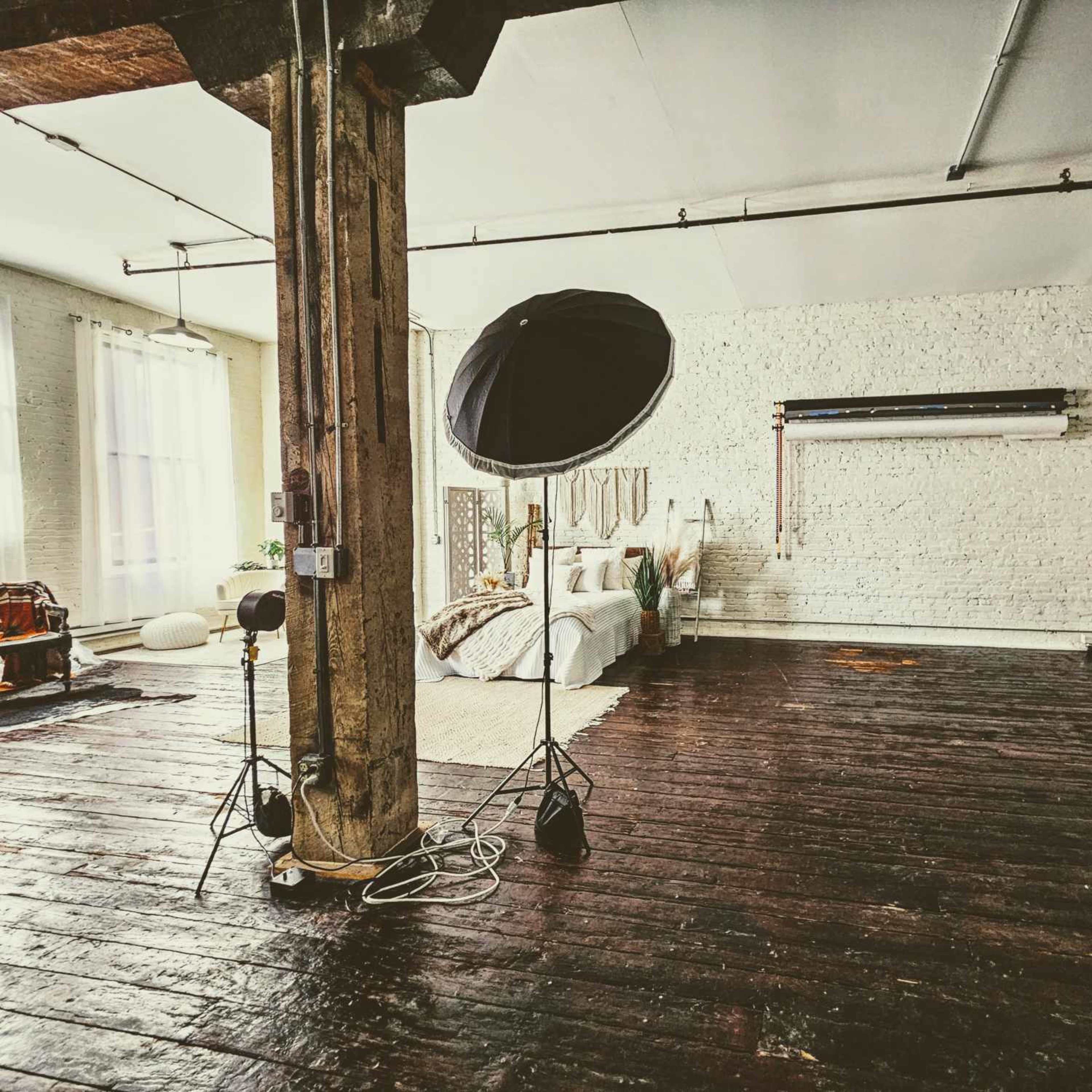 A spacious, well-lit bedroom with wooden floors, a large bed, and a photography setup including a softbox and a backdrop.