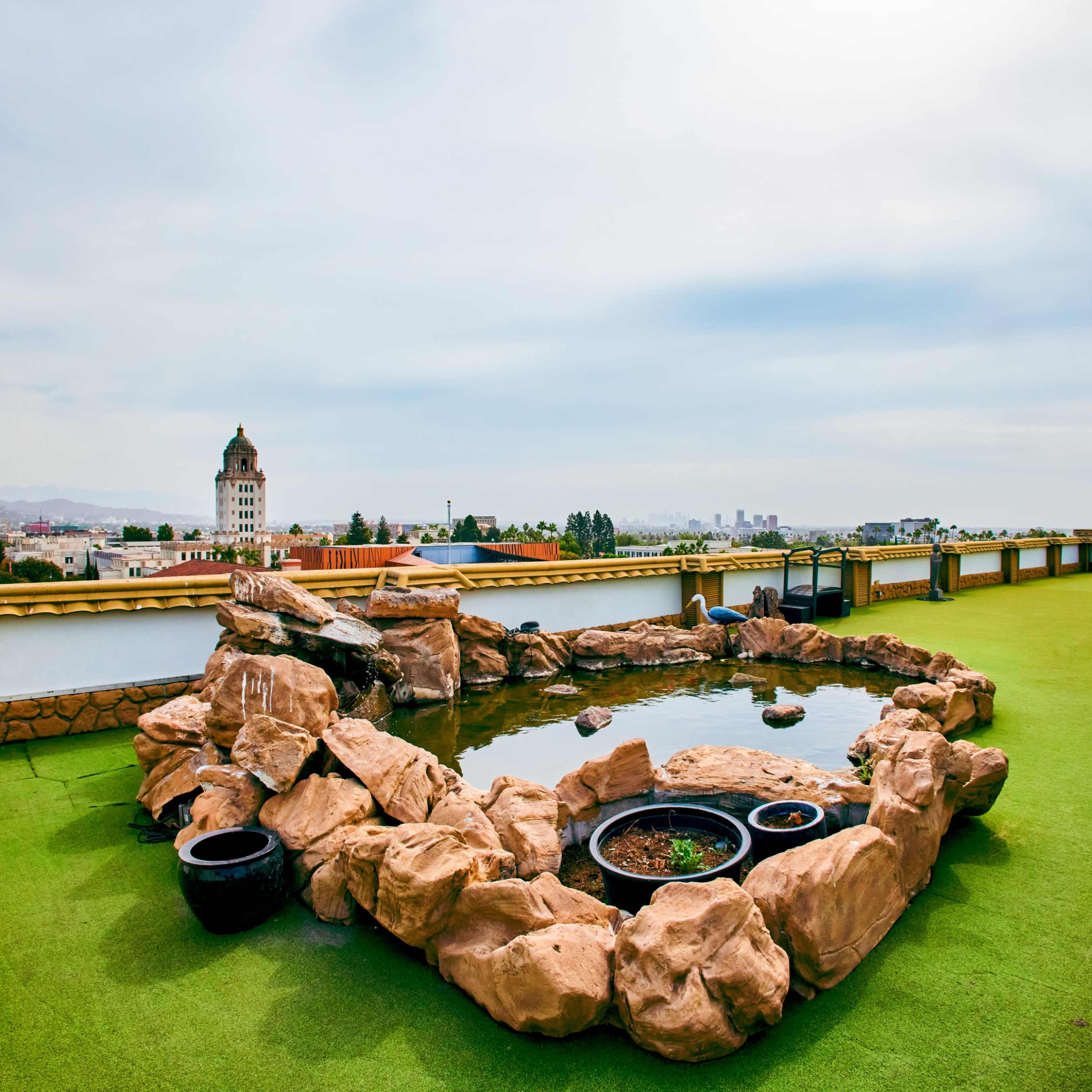 A rooftop terrace featuring a rock formation surrounding a pond, with a view of a cityscape in the background.