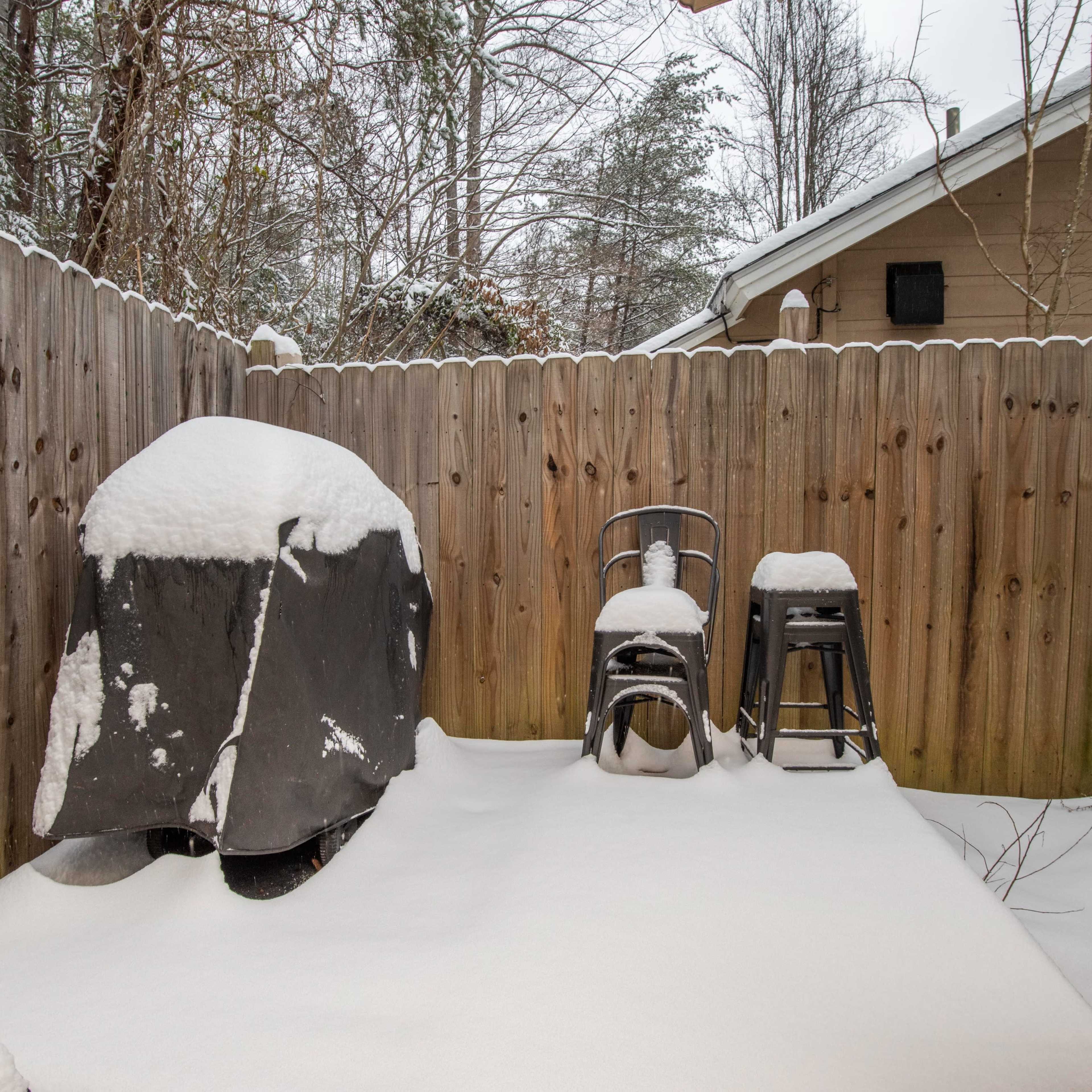 A covered grill and two stools are visible in a snowy backyard surrounded by a wooden fence.