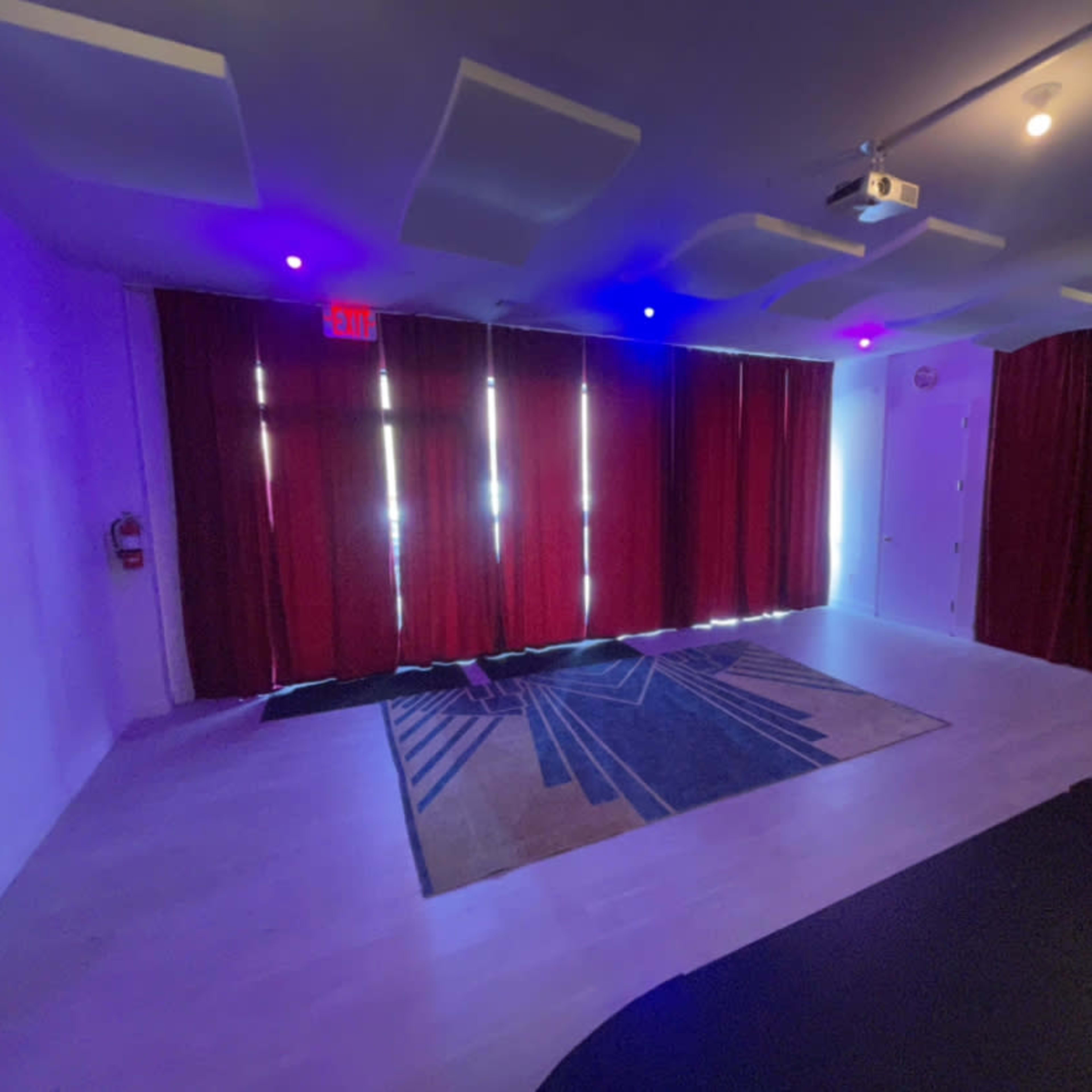 A spacious room with wooden flooring, a patterned area rug, red curtains, and overhead lighting.