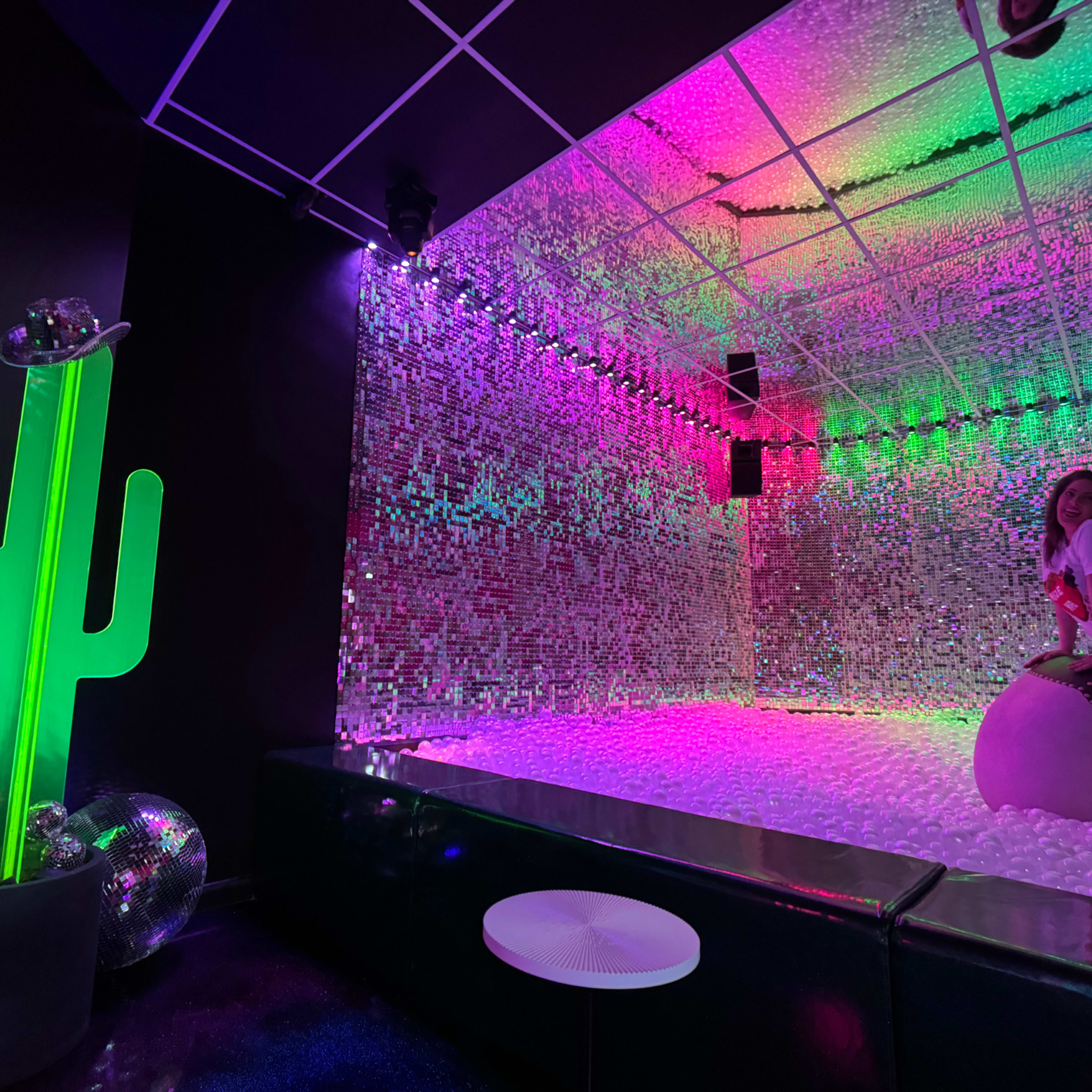 The scene features a brightly colored room with a mirrored wall, a large cactus sculpture, and a woman sitting on a large inflatable object surrounded by a ball pit.