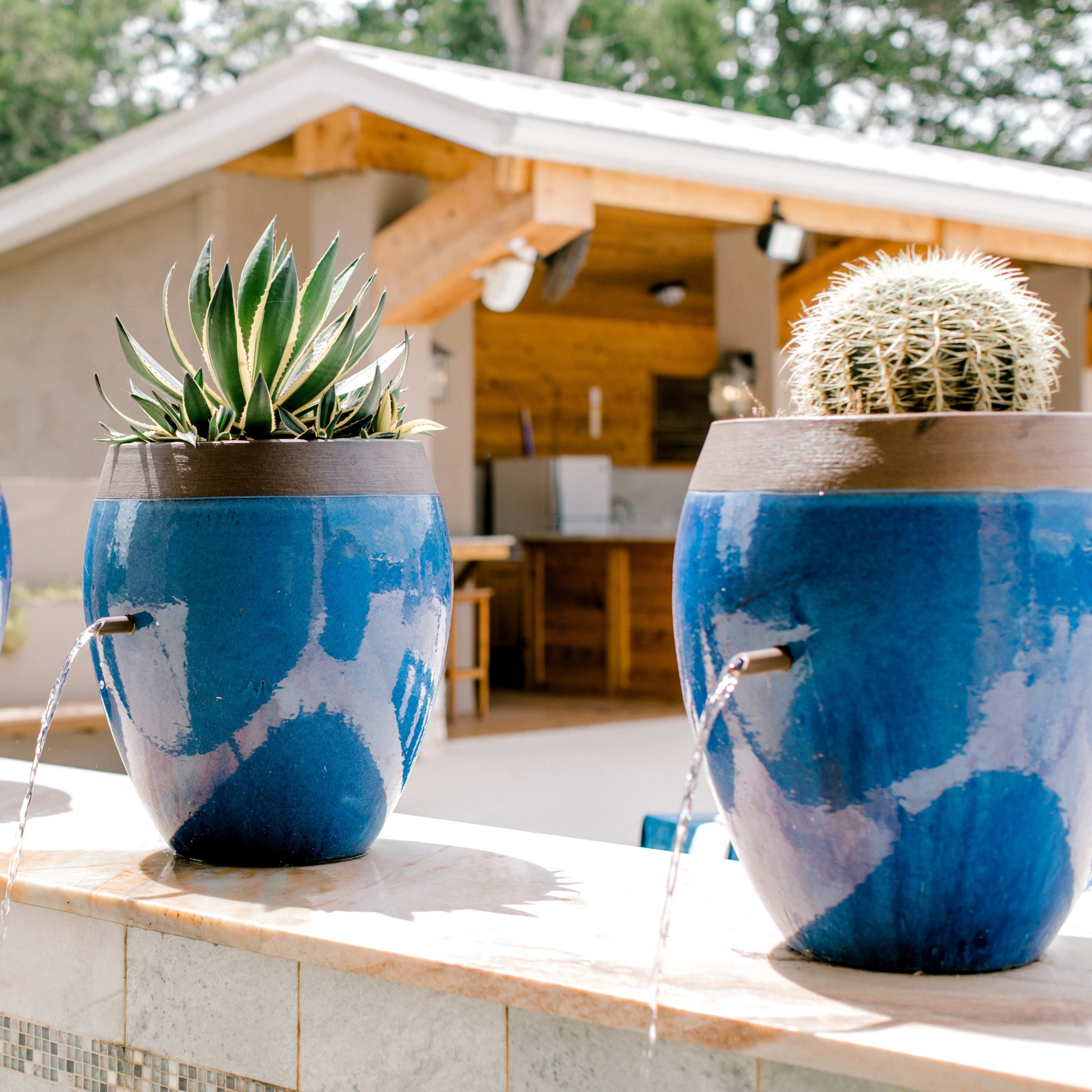 Three blue ceramic pots with cacti and succulents are placed on a stone wall, each featuring a small water fountain.