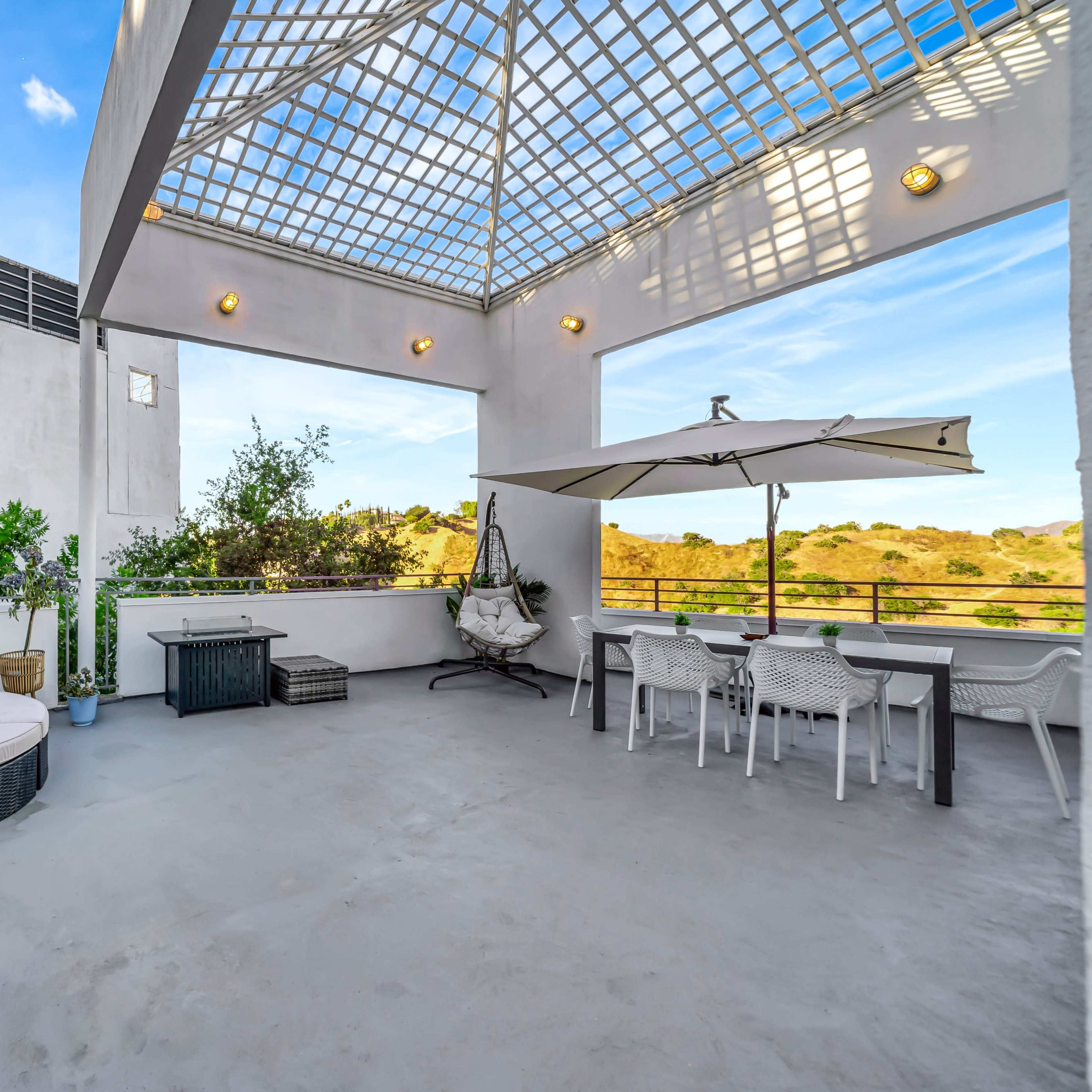 A spacious rooftop terrace featuring a circular lounging area, a dining table under an umbrella, and greenery along the periphery.