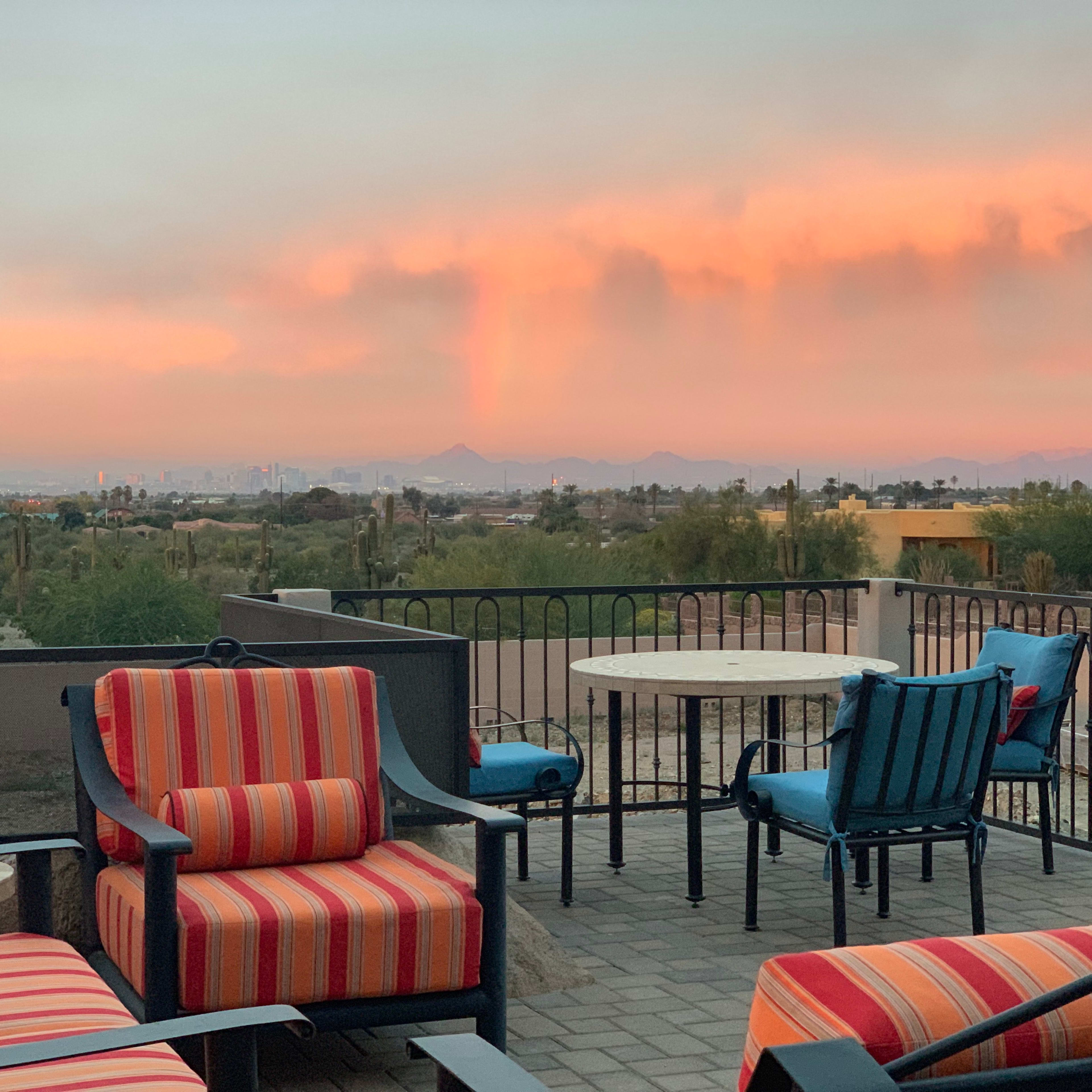 A patio with striped chairs and a table overlooking a sunset-lit landscape featuring distant mountains and a city skyline.