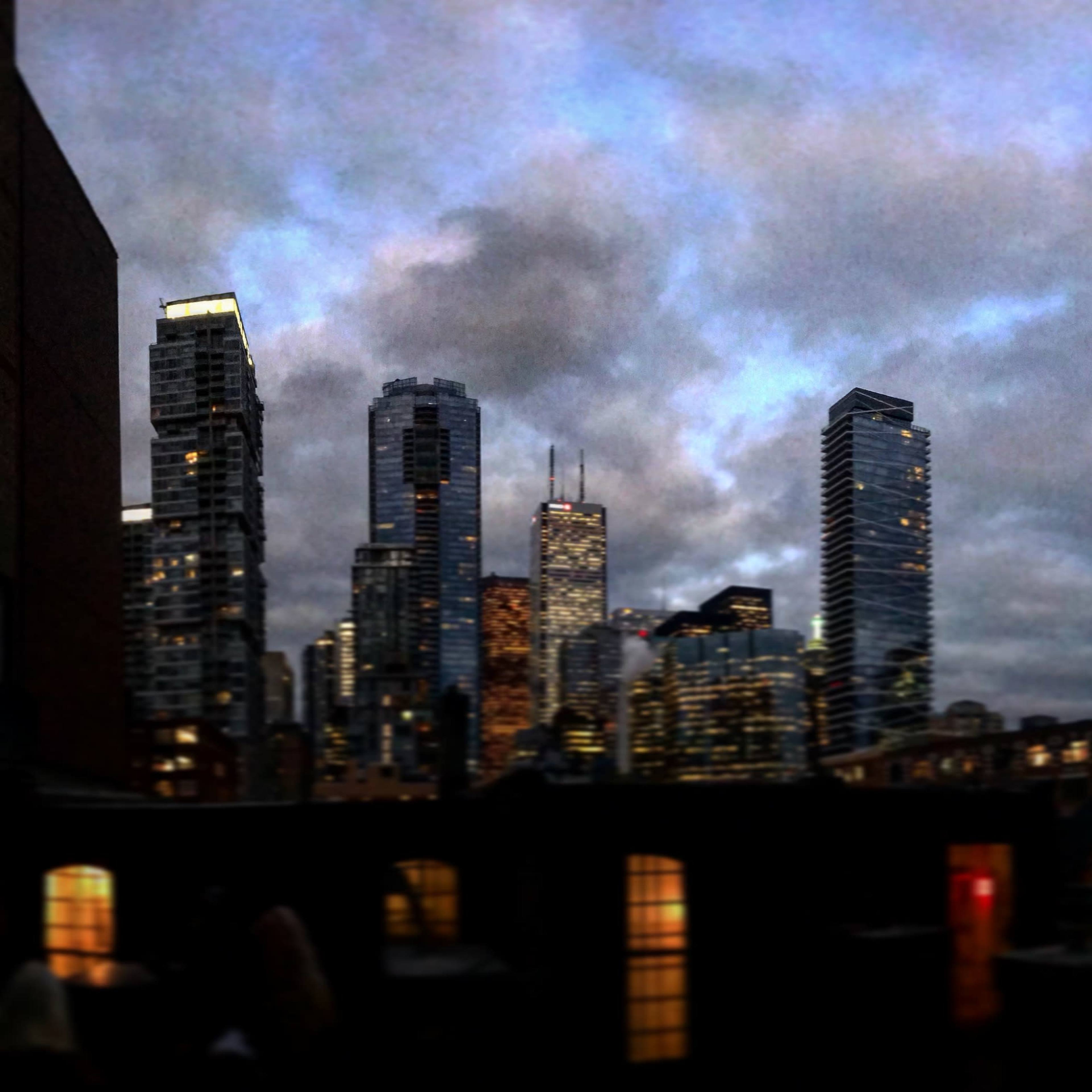 A skyline of tall buildings and skyscrapers in a city at dusk, with a mix of illuminated windows and dark clouds overhead.