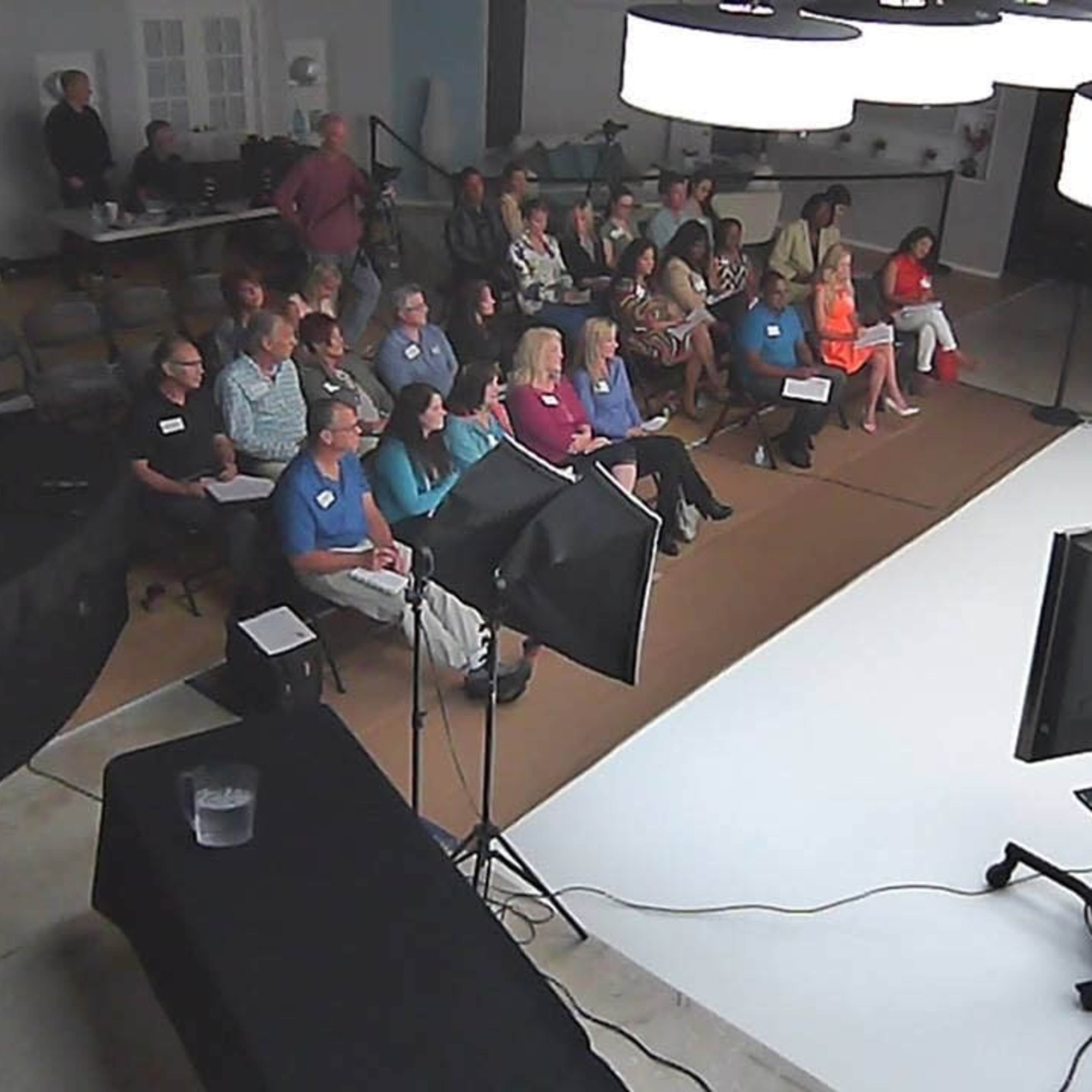 A group of people is seated in a studio audience facing a speaker near a large screen and bright lighting setup.