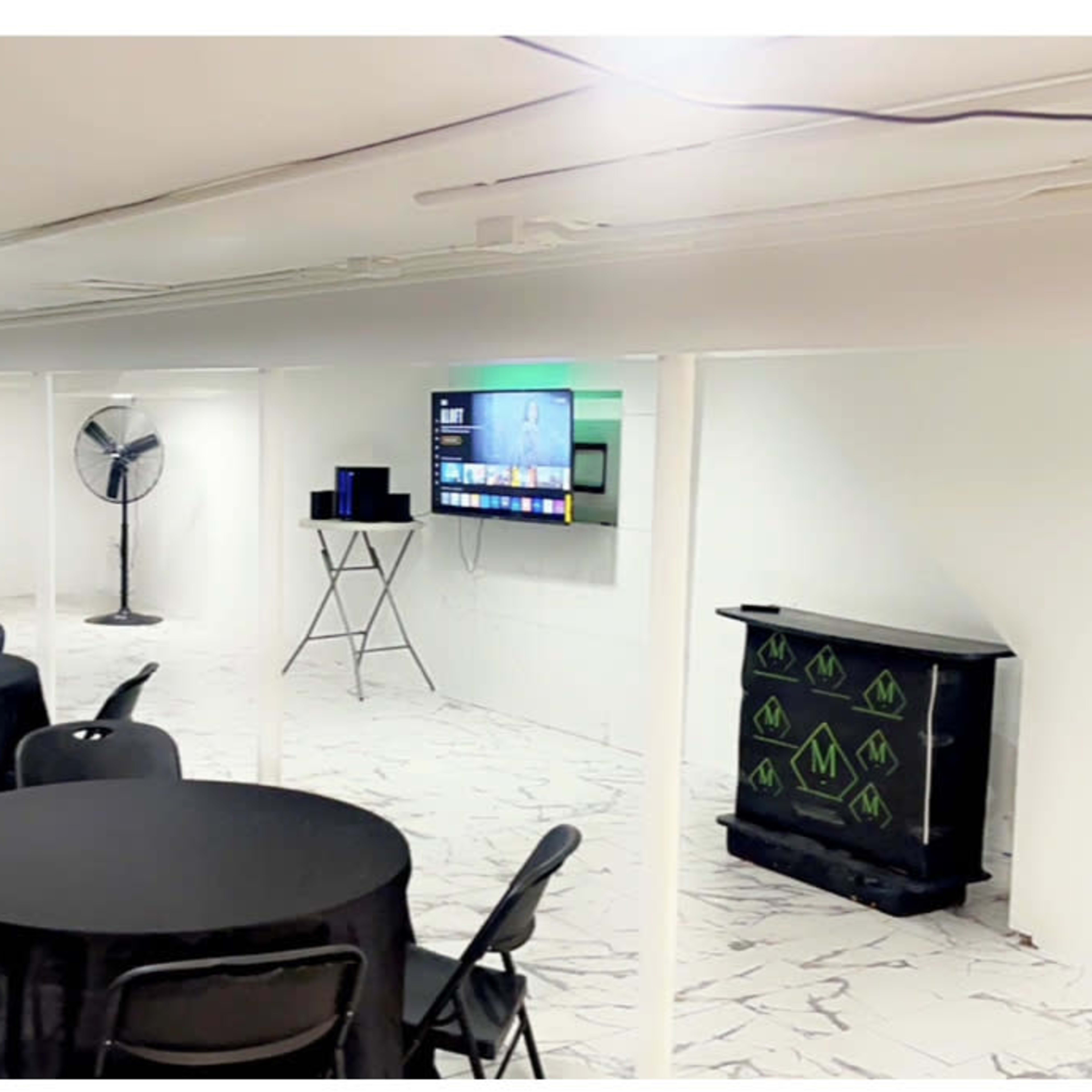 A clean, white-walled meeting space with several black tables and chairs, a fan, and a television mounted on the wall.